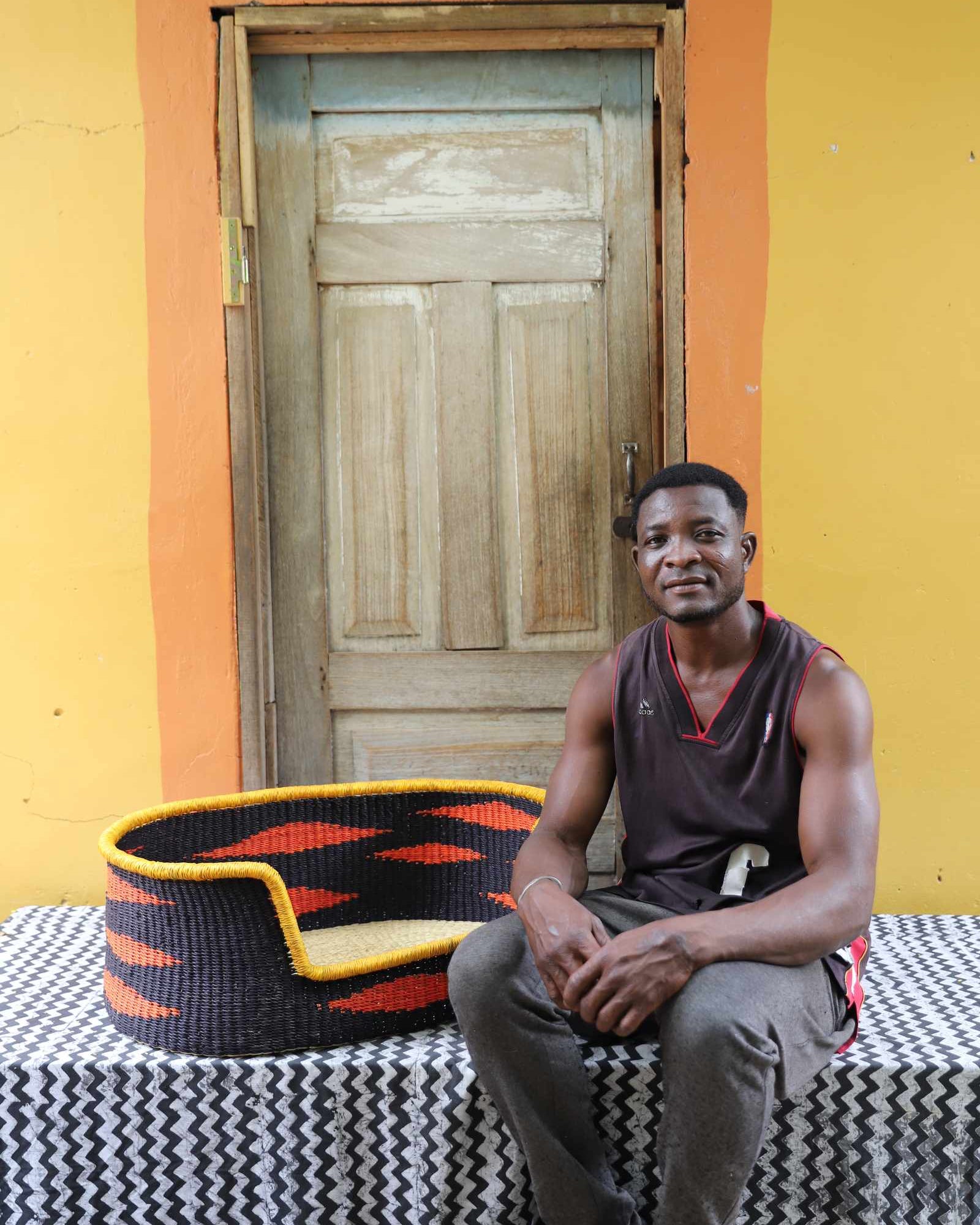 Medium handwoven Bolga pet bed in black with orange flame motif, beside a seated man in a dark vest.