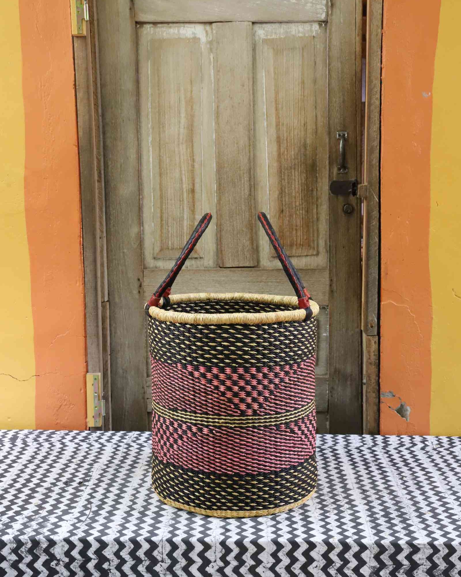 Handwoven open-top Bolga laundry basket in natural straw with black and red woven bands and twin looped handles.