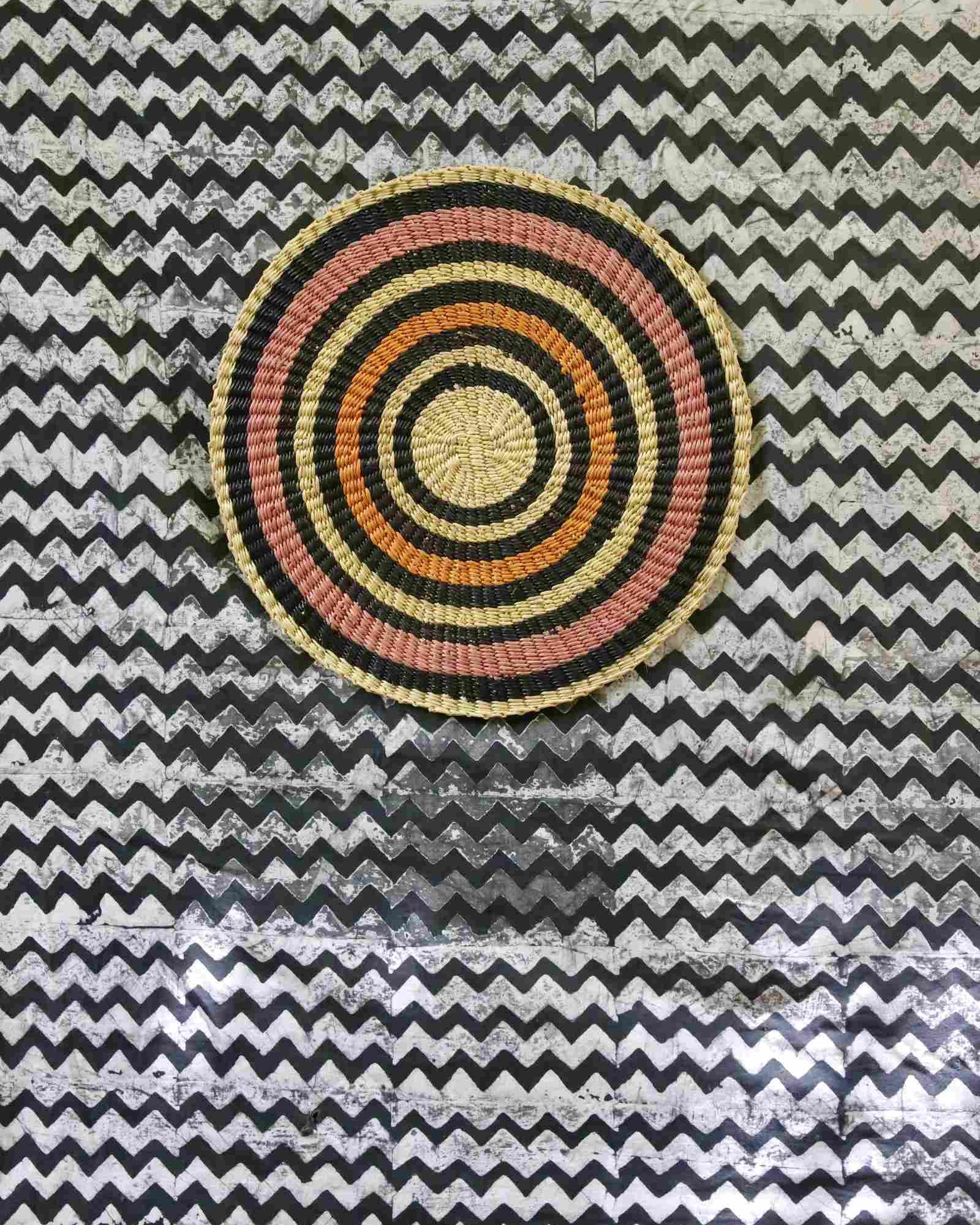 Handwoven Bolga placemat with concentric rings in natural straw, black, orange and pink, on a chevron-print backdrop.