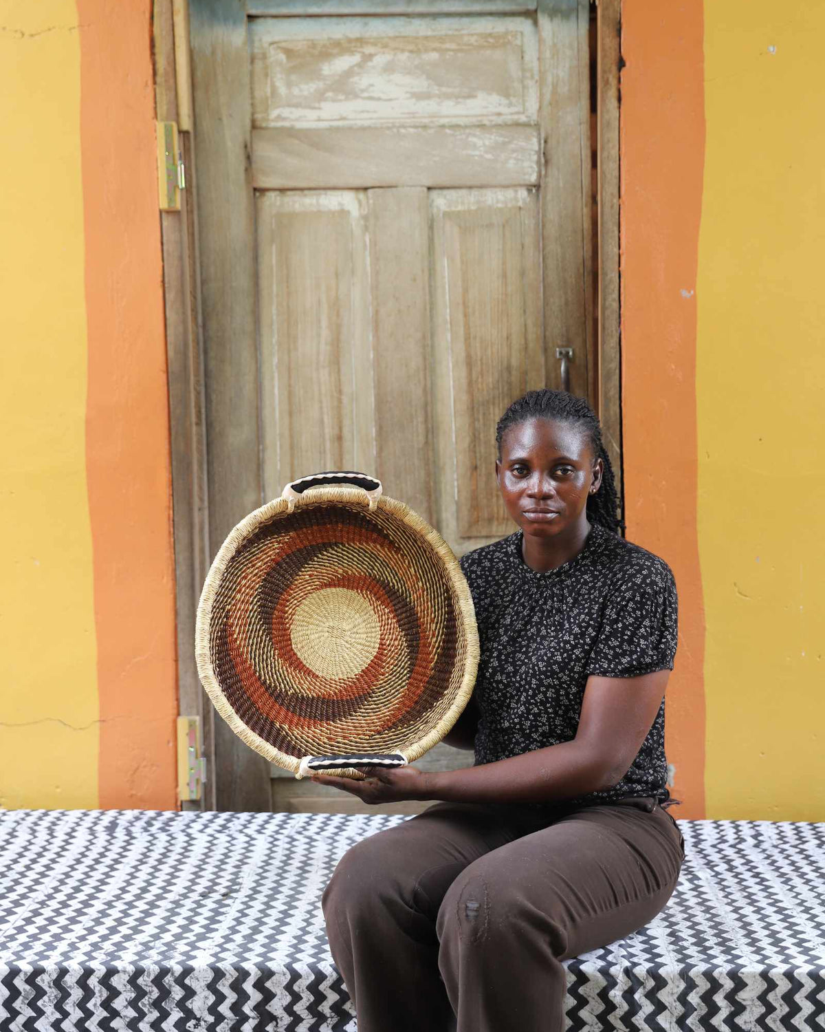 Handwoven Bolga wall tray disc with rust-red pinwheel, held by a seated woman in dark top.