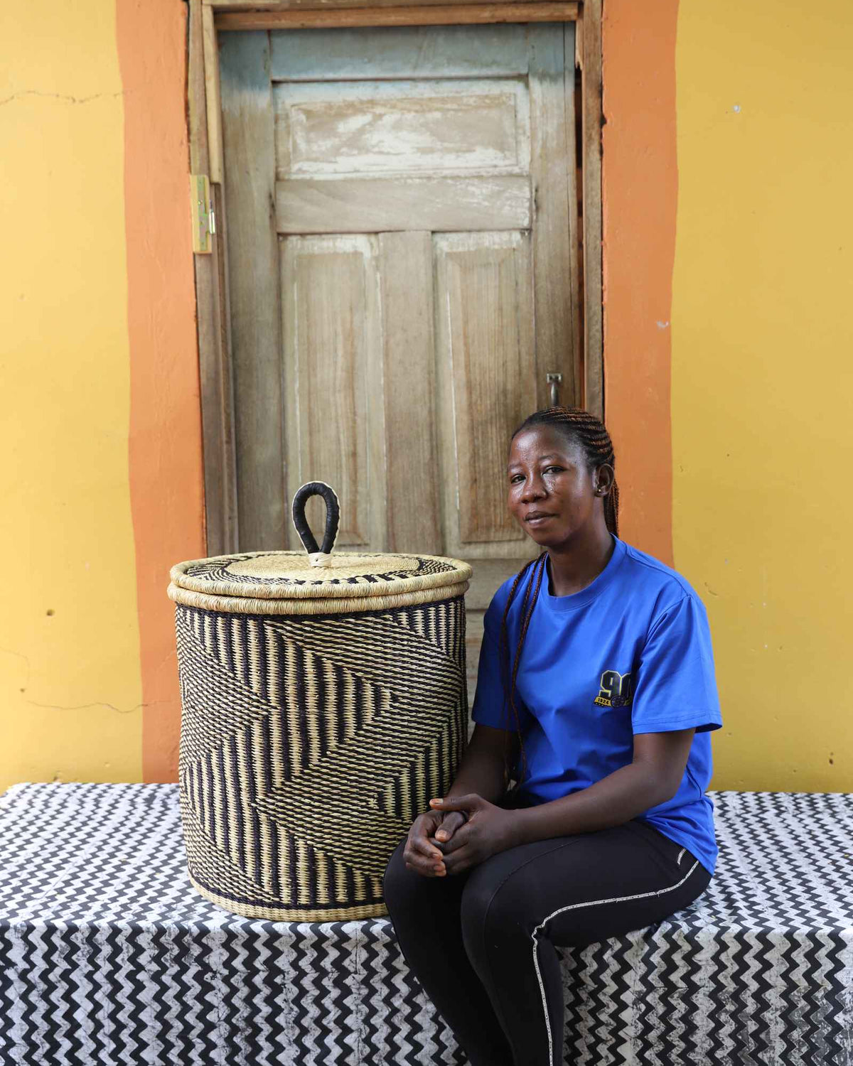 Handwoven Bolga laundry basket in natural with dark wave stripes, beside a seated woman in blue.
