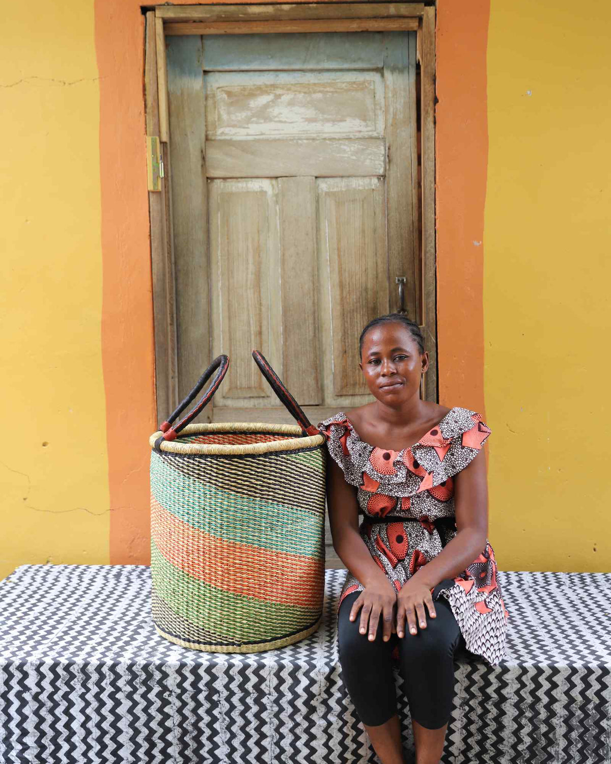 Medium handwoven Bolga laundry basket in green, orange and pink stripes, beside a seated woman in floral.