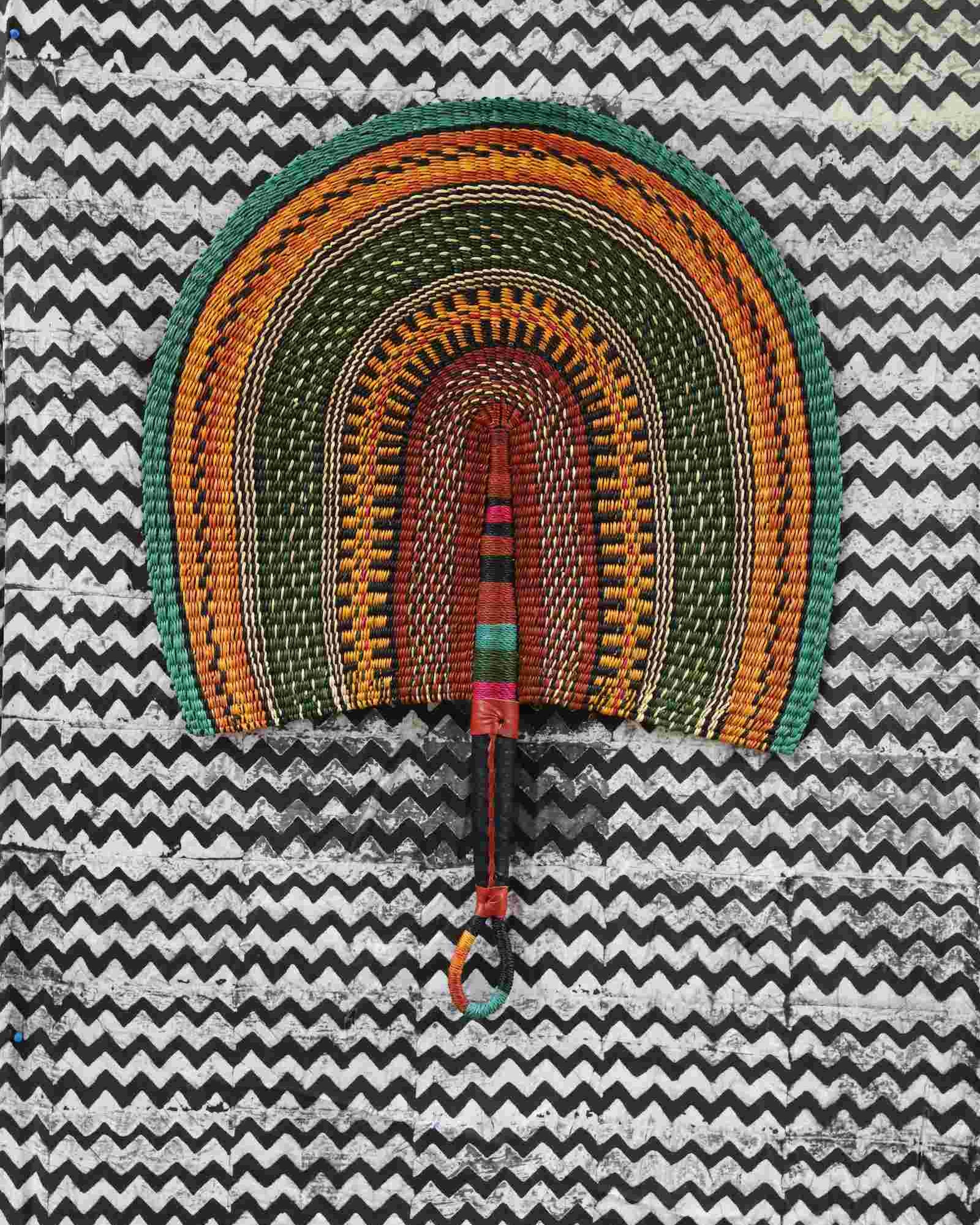 Naba Penga wall fan in natural straw with teal, orange and green concentric bands, laid on a chevron cloth.
