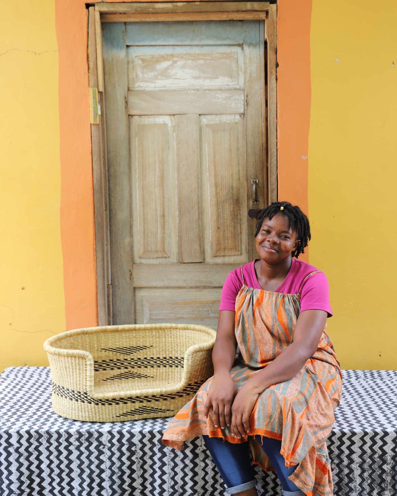 Small handwoven Bolga pet bed in natural straw with subtle black motif, beside a seated woman in pink.