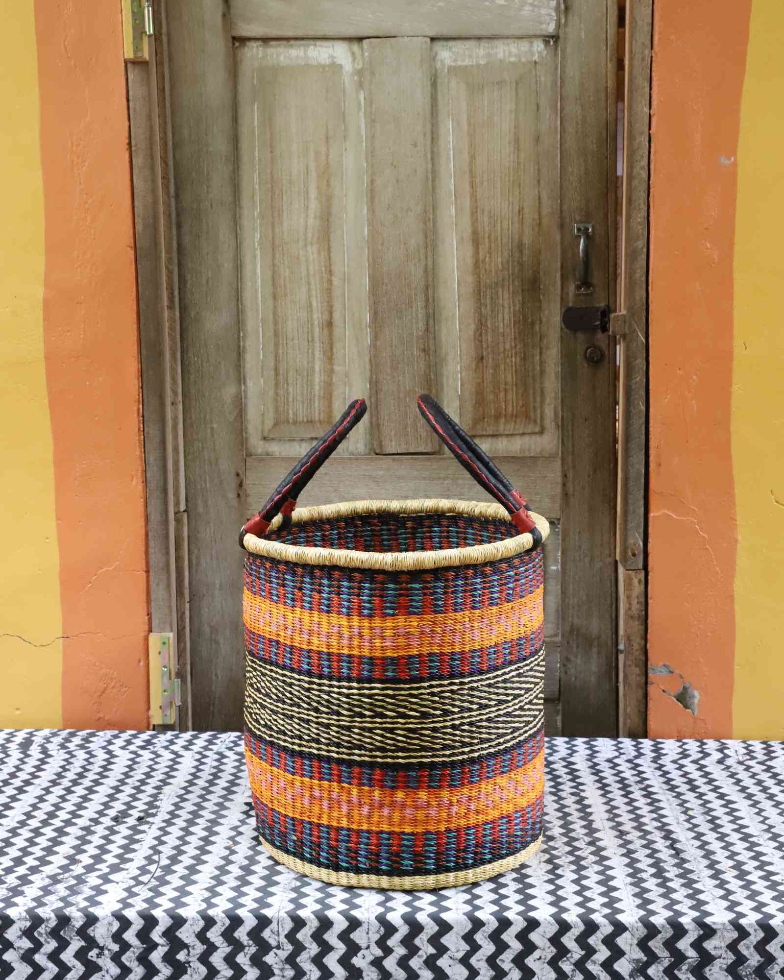 Handwoven open-top Bolga laundry basket in natural straw with orange, yellow and black stripes and twin handles.
