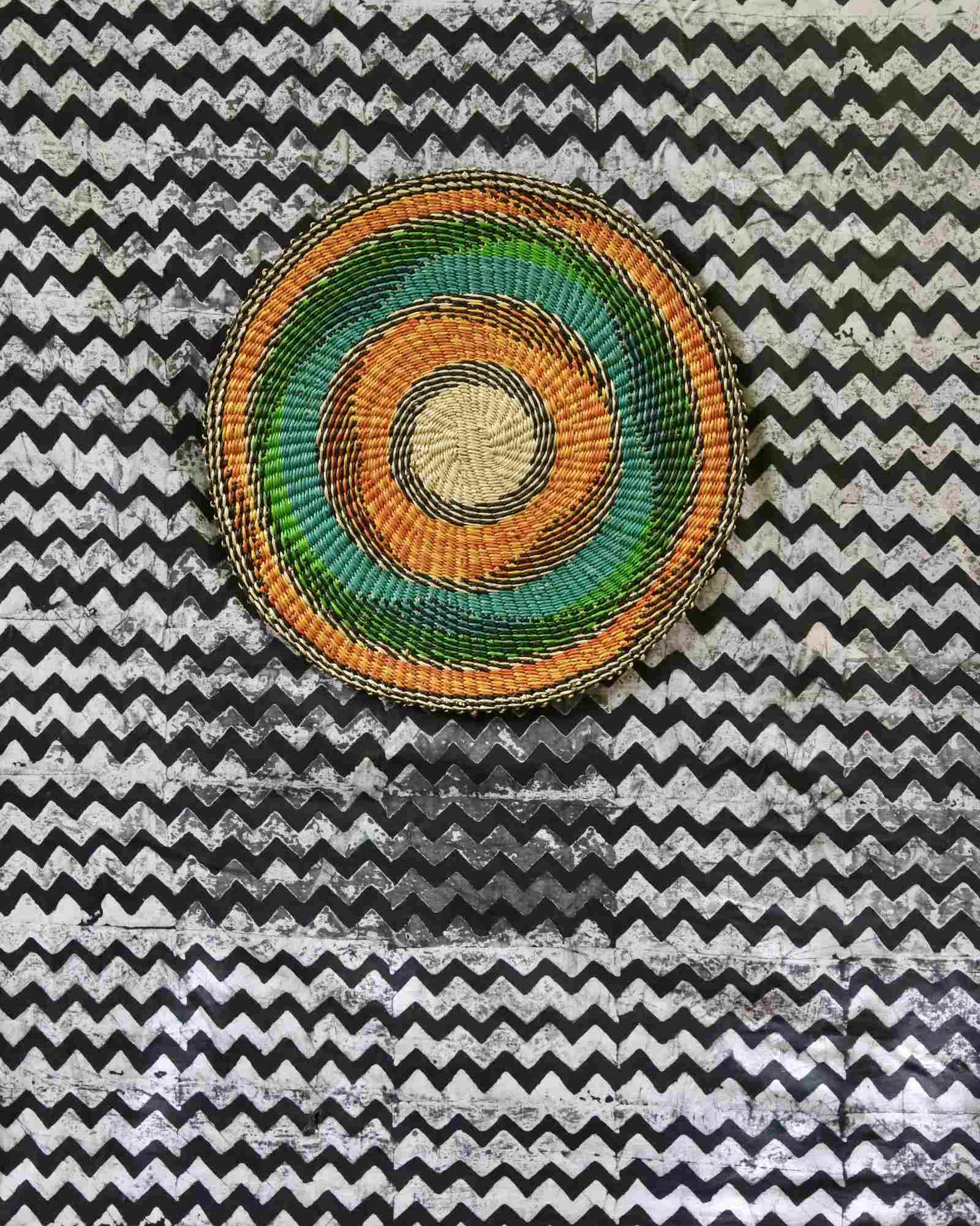 Handwoven Bolga placemat with concentric rings in natural straw, orange, teal and green, on a chevron-print backdrop.