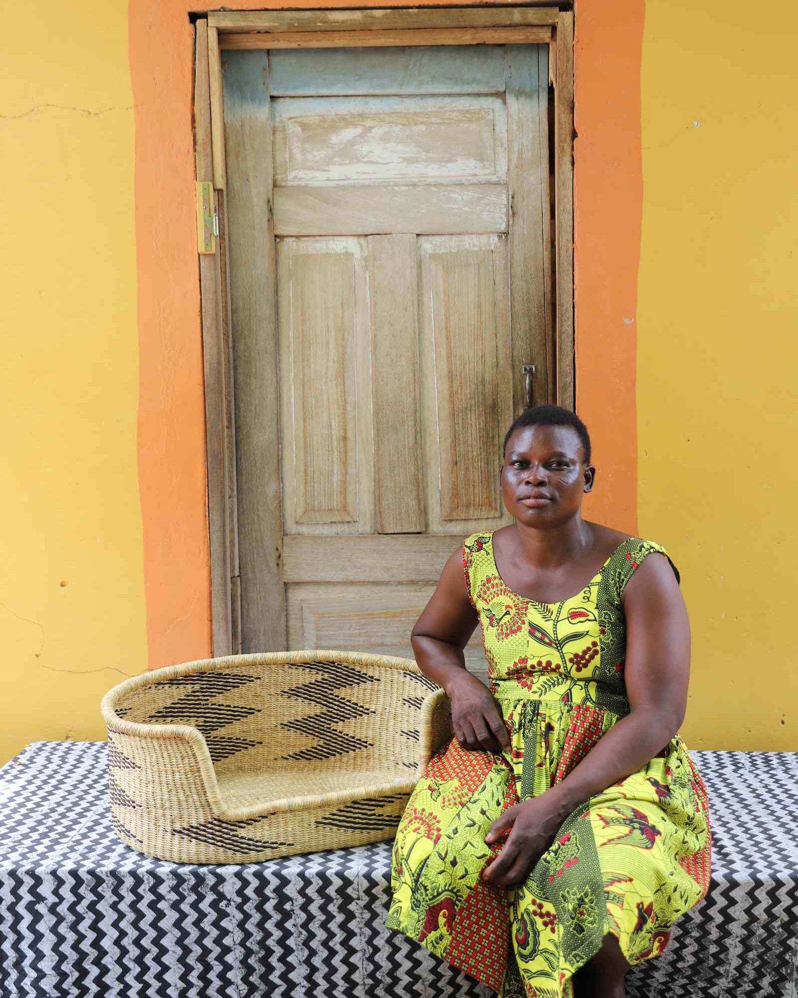 Small handwoven Bolga pet bed in natural straw with black arrow pattern, beside a seated woman in yellow.