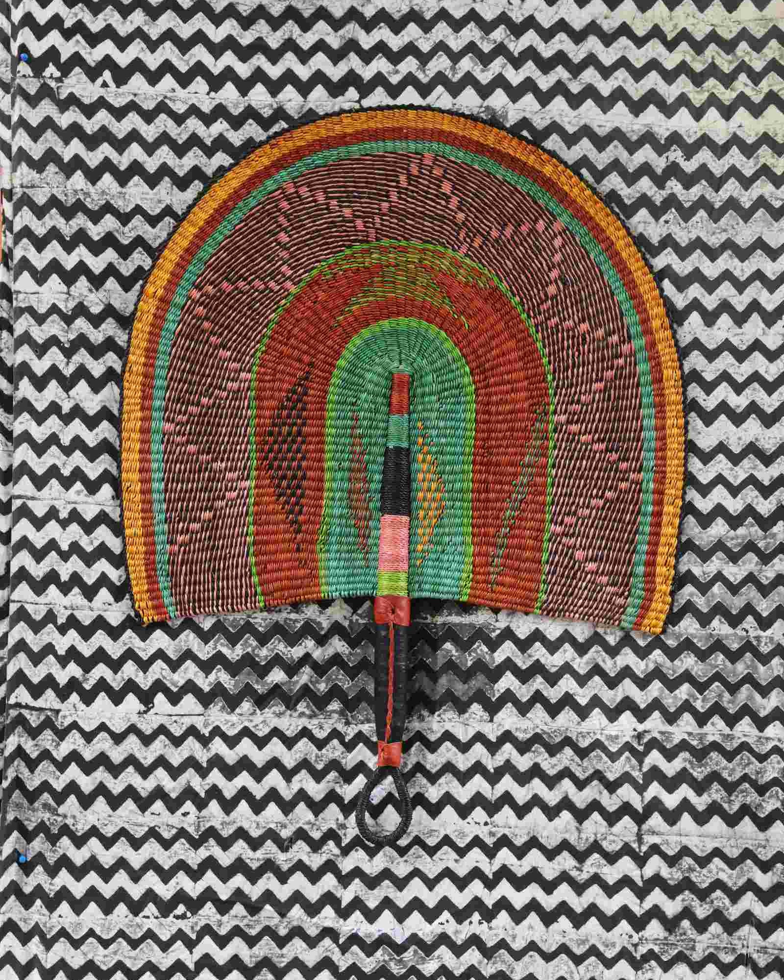 Naba Penga wall fan in natural straw with green, orange and black concentric bands, laid on a chevron cloth.