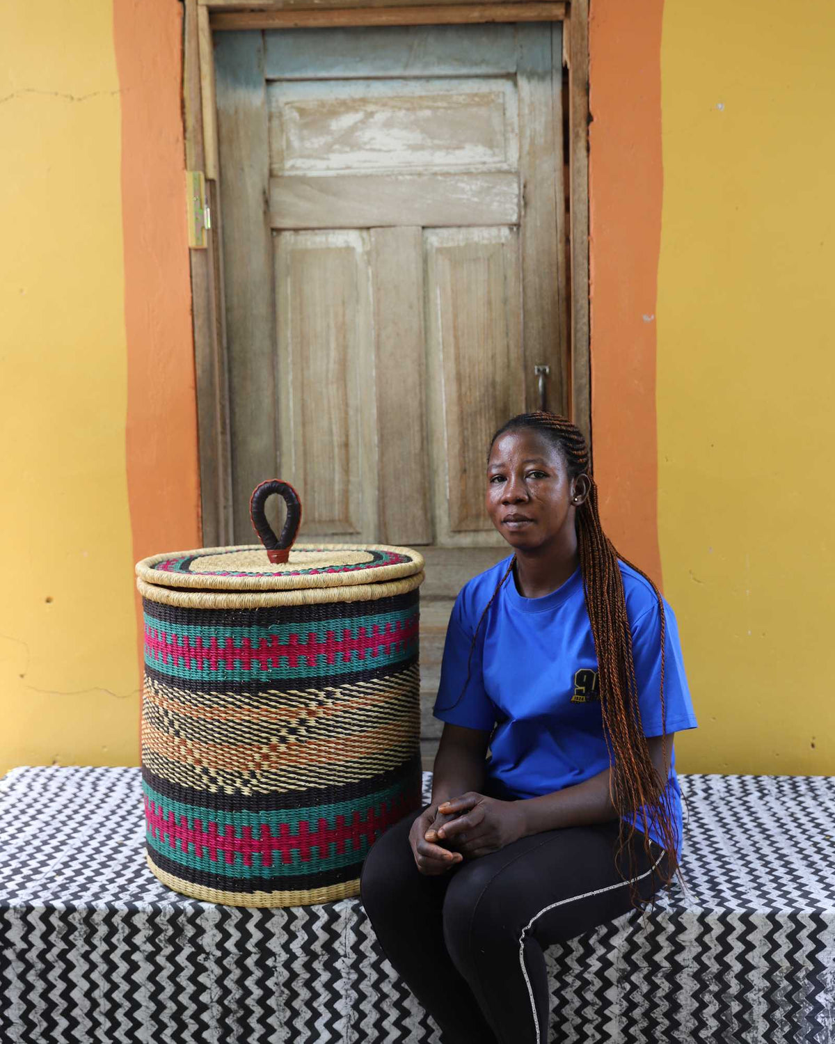 Handwoven Bolga laundry basket in fine pink, black and orange bands, beside a seated woman in blue.