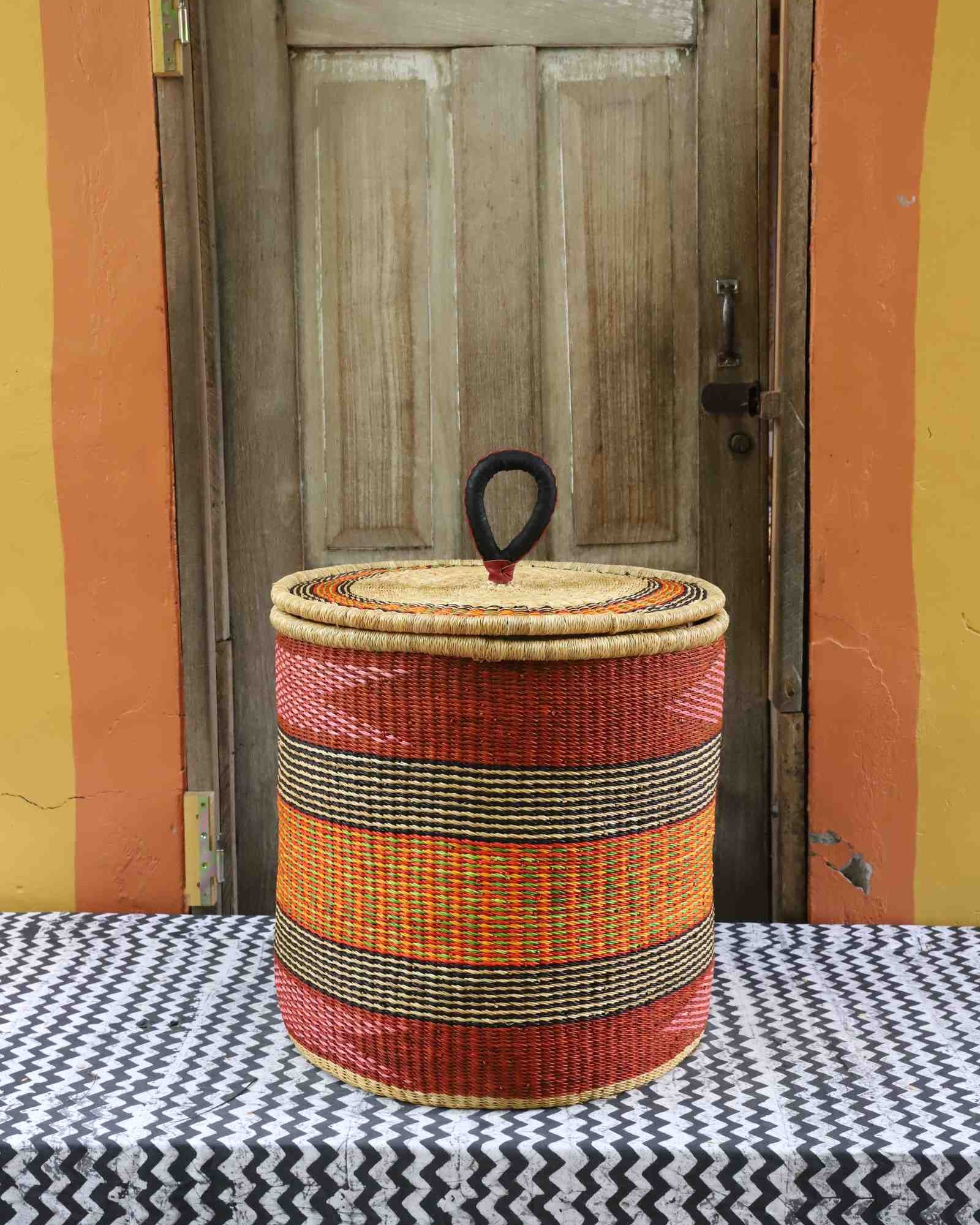 Handwoven Bolga laundry basket with coiled rim and lid in deep red with orange and yellow banded stripes.
