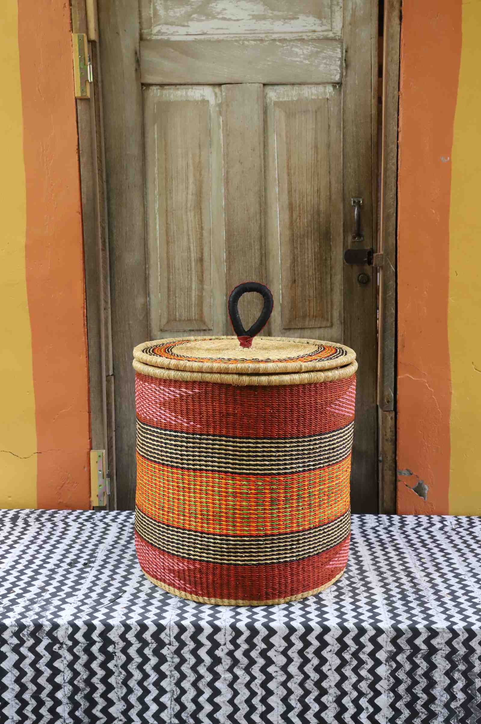 Handwoven Bolga laundry basket with coiled rim and lid in deep red with orange and yellow banded stripes.
