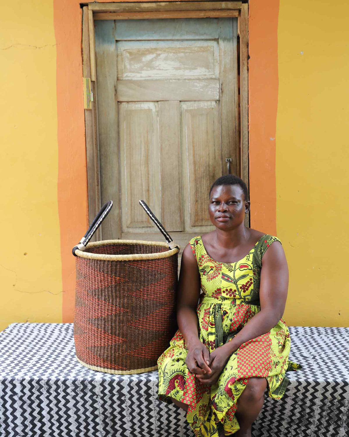 Medium handwoven Bolga laundry basket in rust-orange with dark zigzag, beside a seated woman in floral.