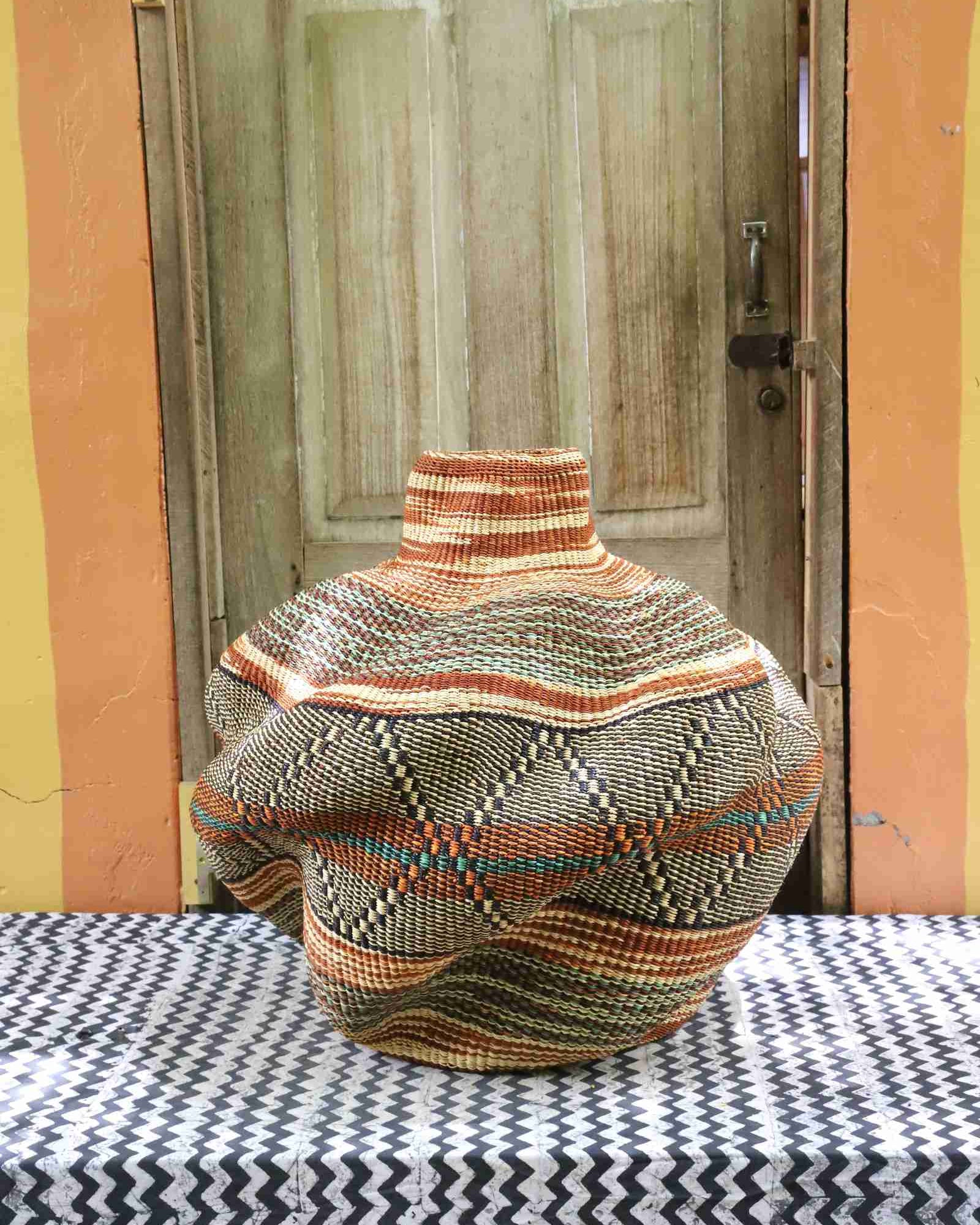 Rear view of a handwoven African art basket showing its sculptural lobed form and earth-tone stripes, on a chevron surface.