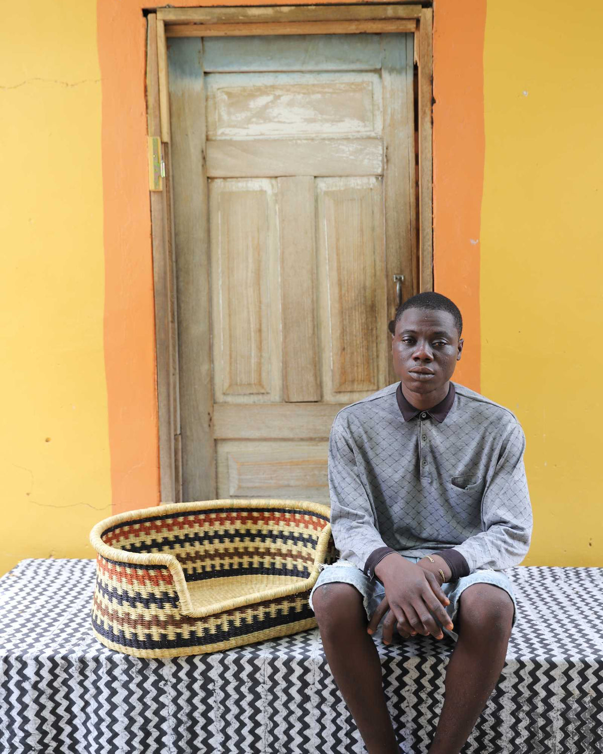 Small handwoven Bolga pet bed in natural straw with black zigzag, beside a seated man in grey.