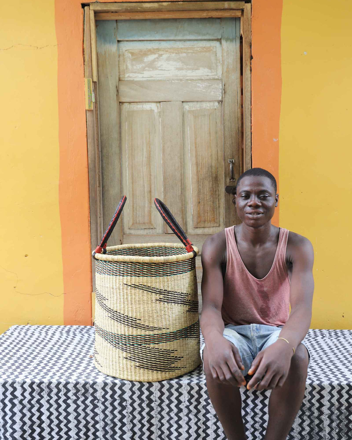 Medium handwoven Bolga laundry basket in natural with scattered black waves, beside a seated man in coral.
