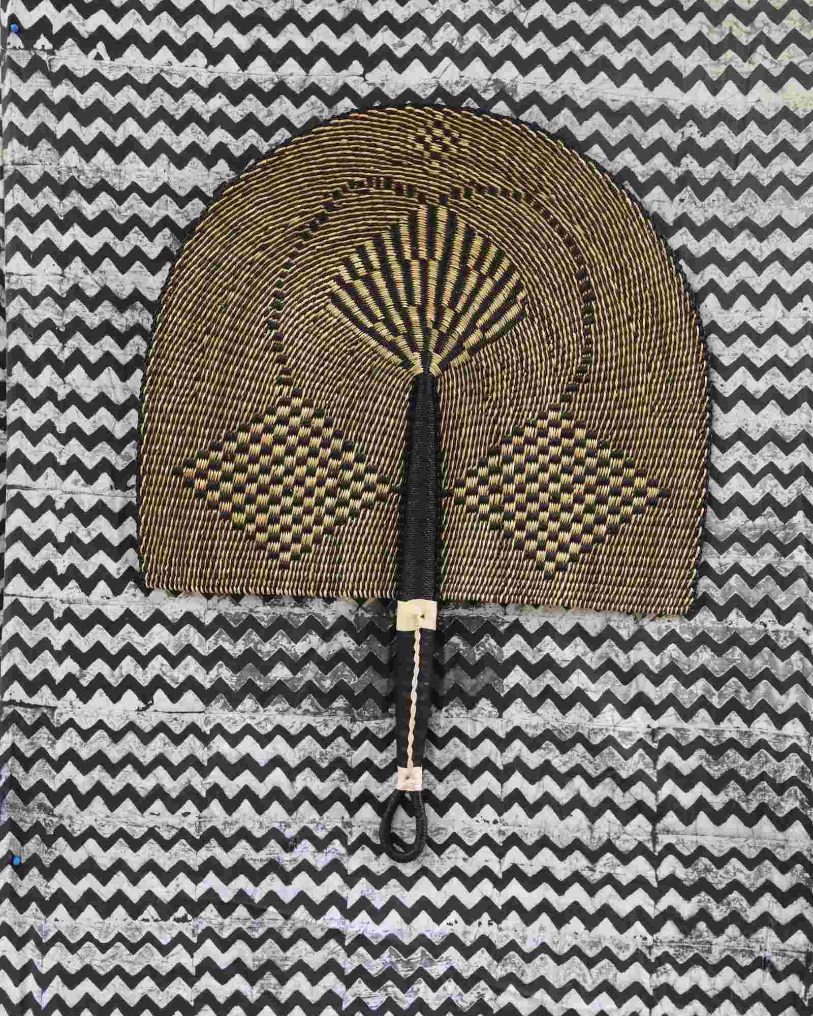 Naba Penga wall fan in natural straw with a dark geometric diamond weave, laid on a chevron cloth.