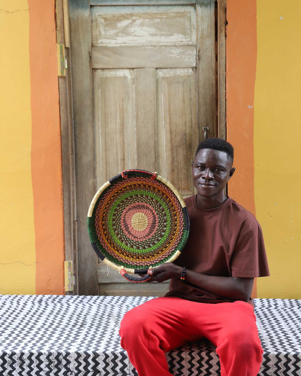Handwoven Bolga wall tray disc with red and green bands, held by a seated man in a brown top.