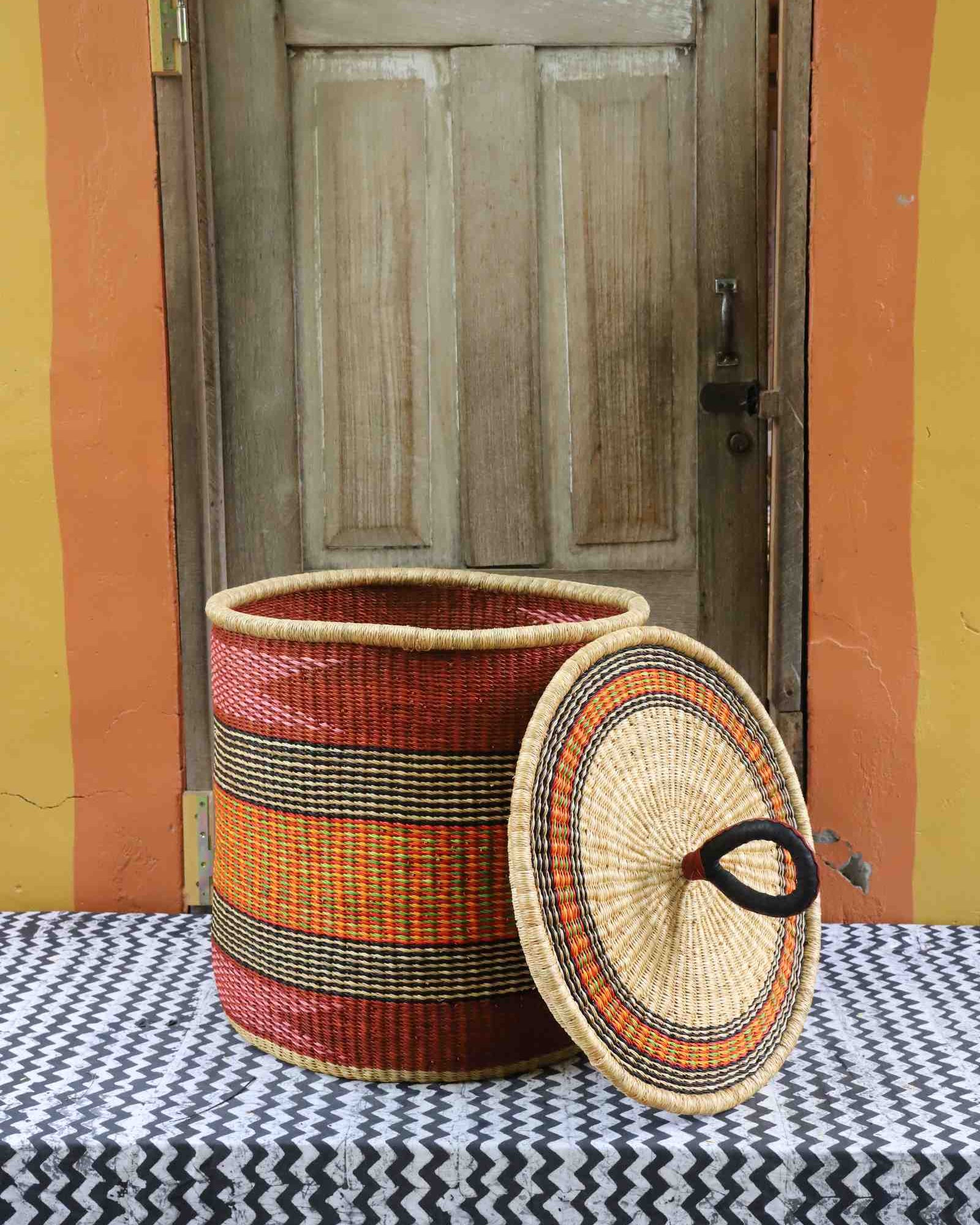 Handwoven Bolga laundry basket with lid removed, deep red body with orange and yellow banded stripes.