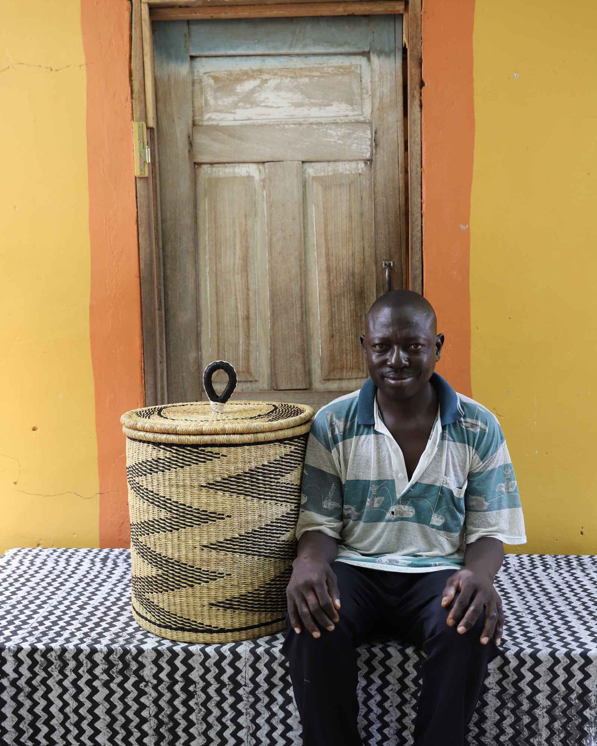 Handwoven Bolga laundry basket in natural straw with black wave motif, beside a seated man in stripes.