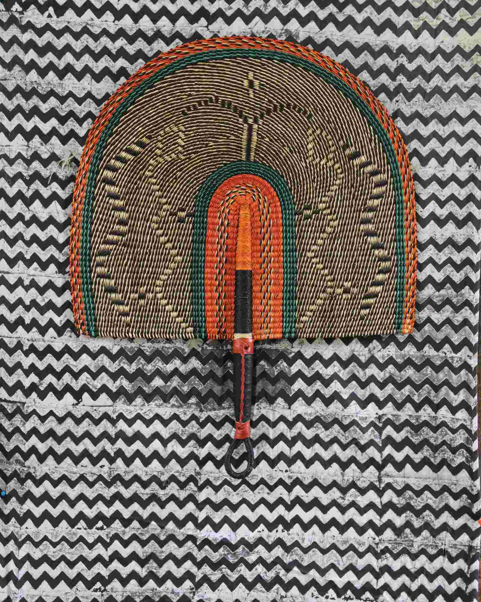 Naba Penga wall fan in natural straw with an orange and green banded border, laid on a chevron cloth.