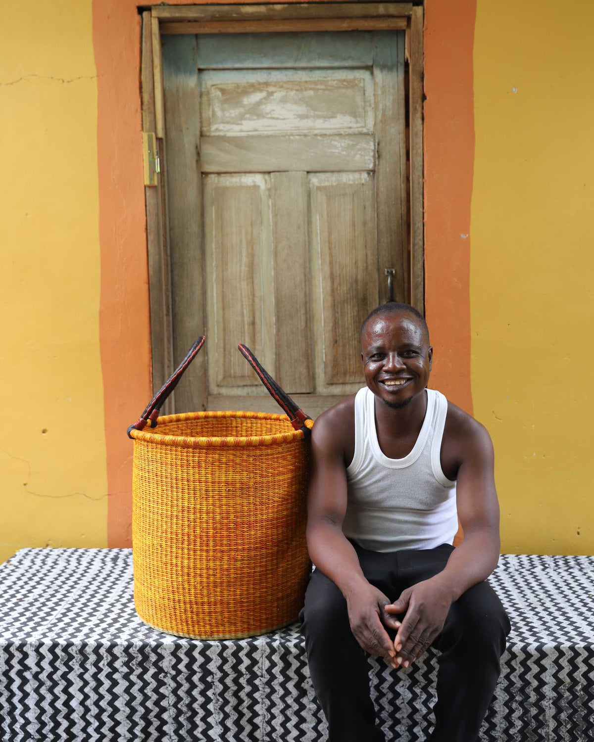 Medium handwoven Bolga laundry basket in dyed bold orange, beside a seated man in a white vest.