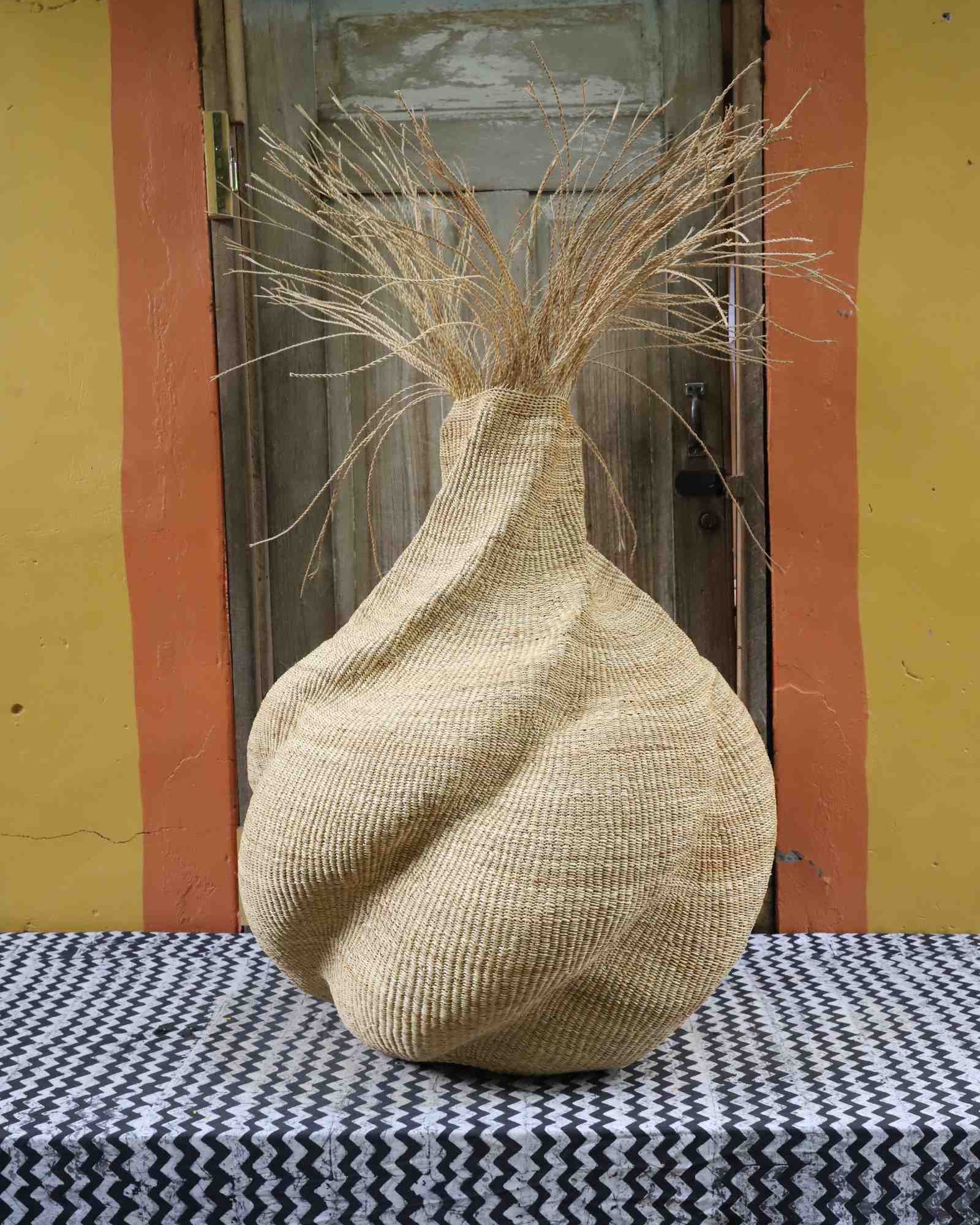 Handwoven art basket in natural straw with sculptural twisted body and dried grass stems sprouting from the top.