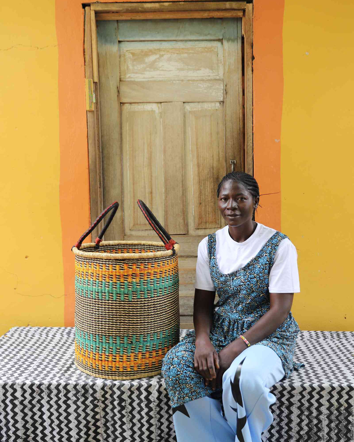 Medium handwoven Bolga laundry basket in natural with orange and teal bands, beside a seated woman in blue.