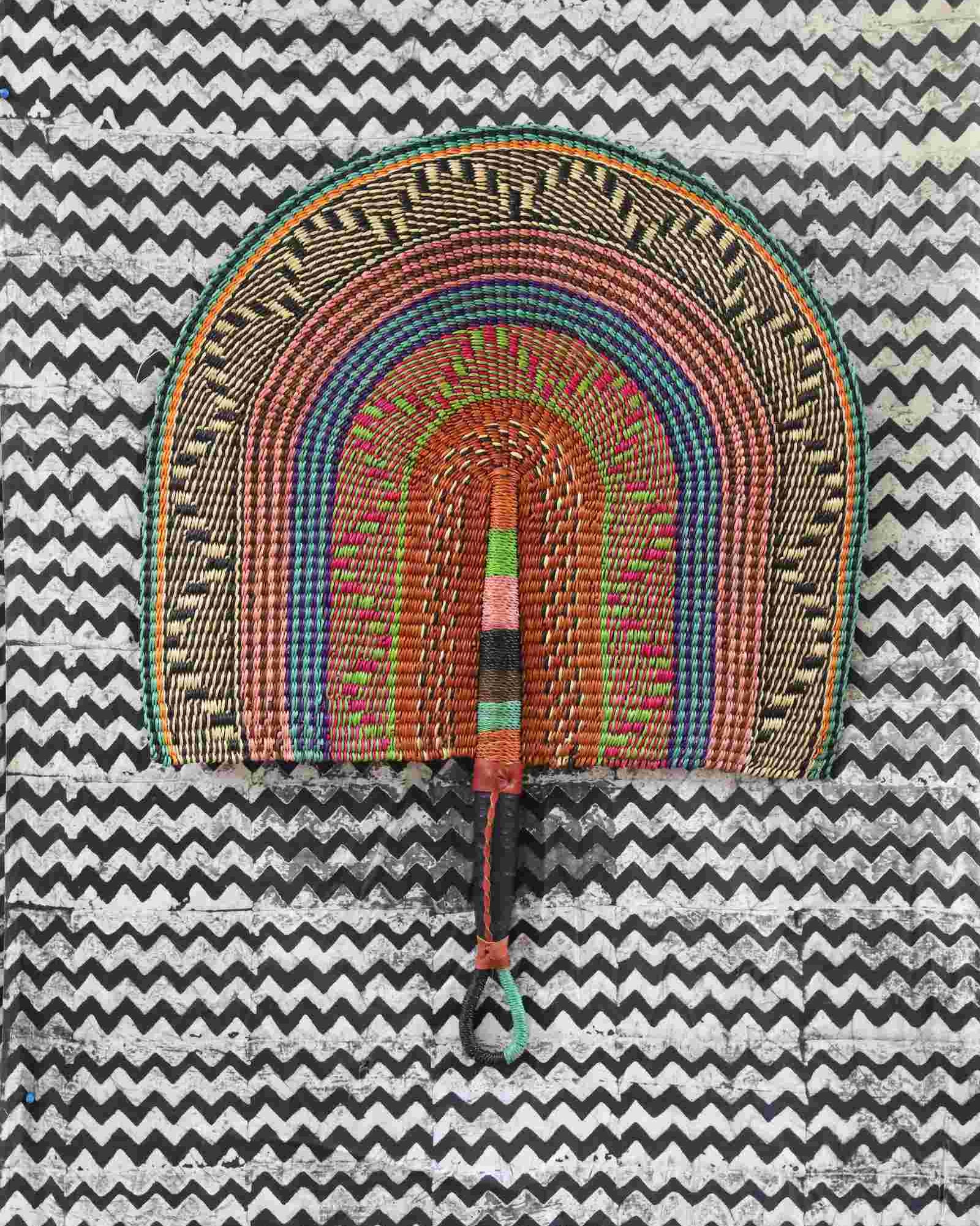 Naba Penga wall fan in natural straw with teal, rust and green multicolour bands, laid on a chevron cloth.