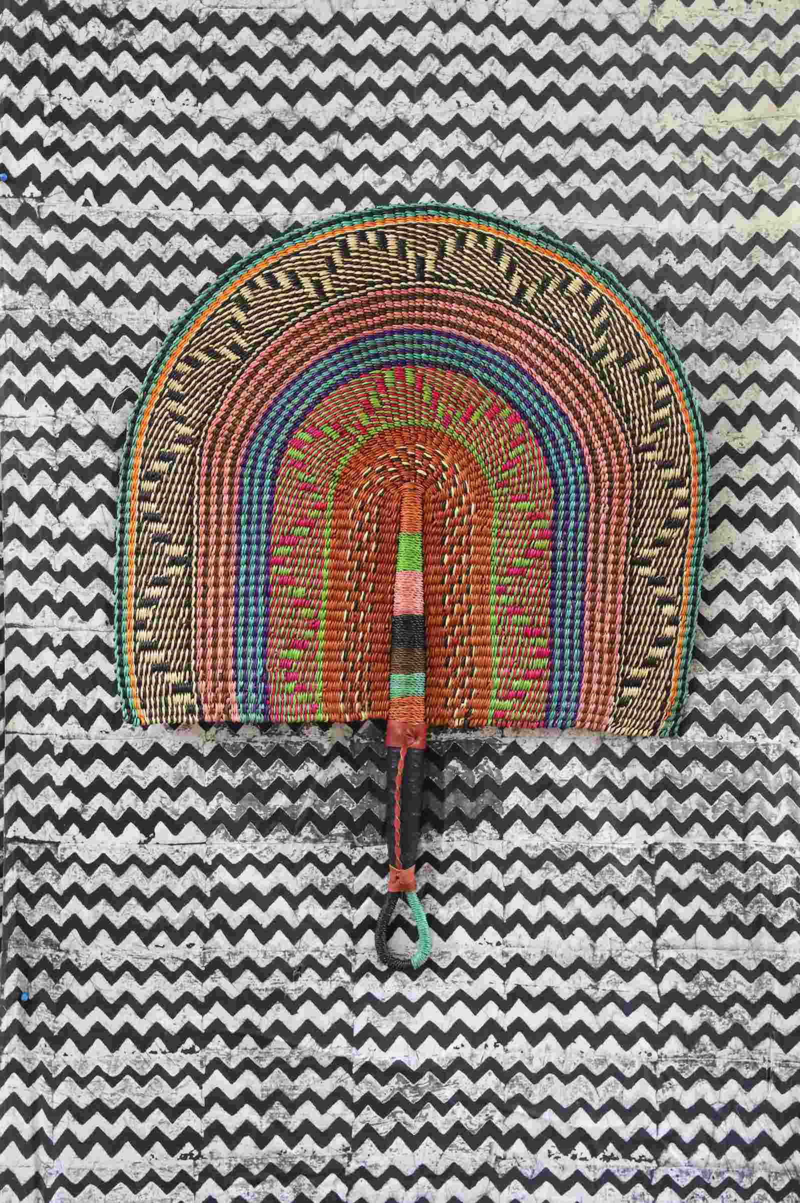 Naba Penga wall fan in natural straw with teal, rust and green multicolour bands, laid on a chevron cloth.