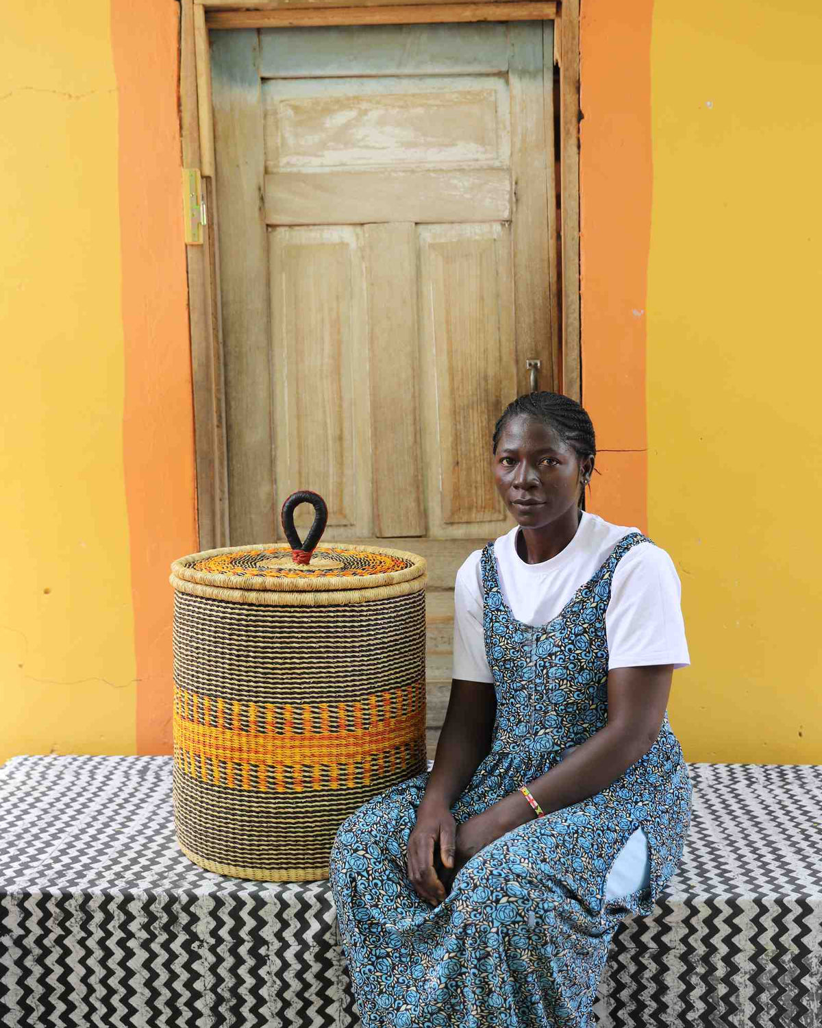 Handwoven Bolga laundry basket in natural straw with orange central band, beside a seated woman in floral.
