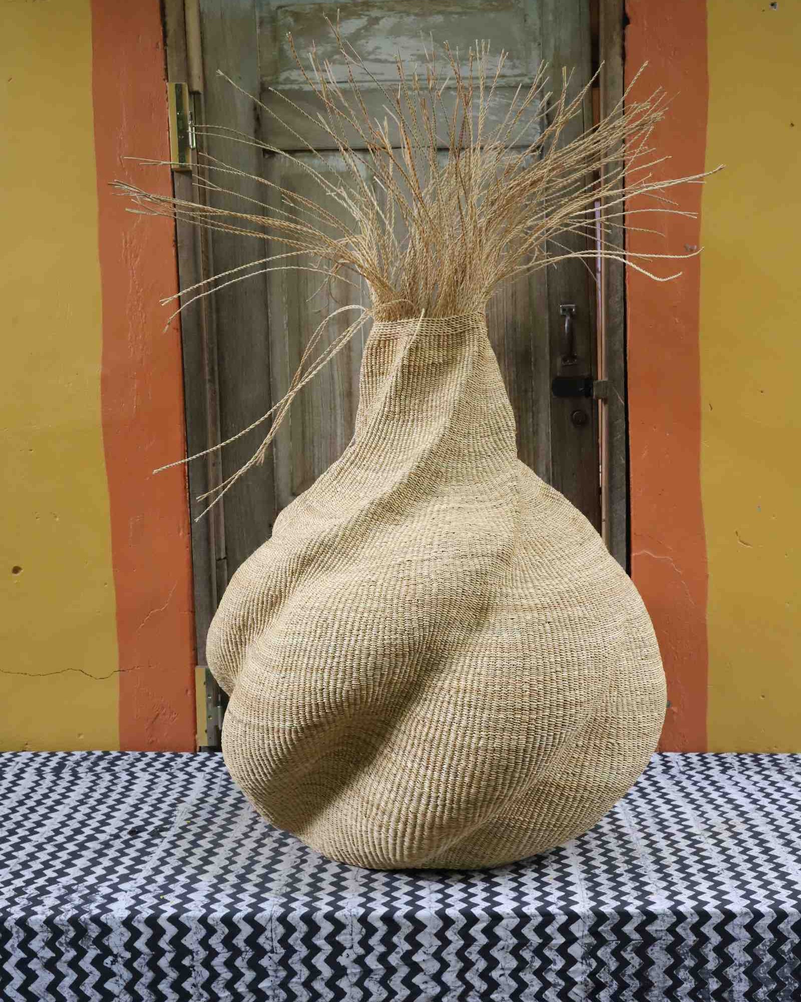 Handwoven art basket in natural straw with sculpted twisted form and dried grass at the top, on a chevron surface.