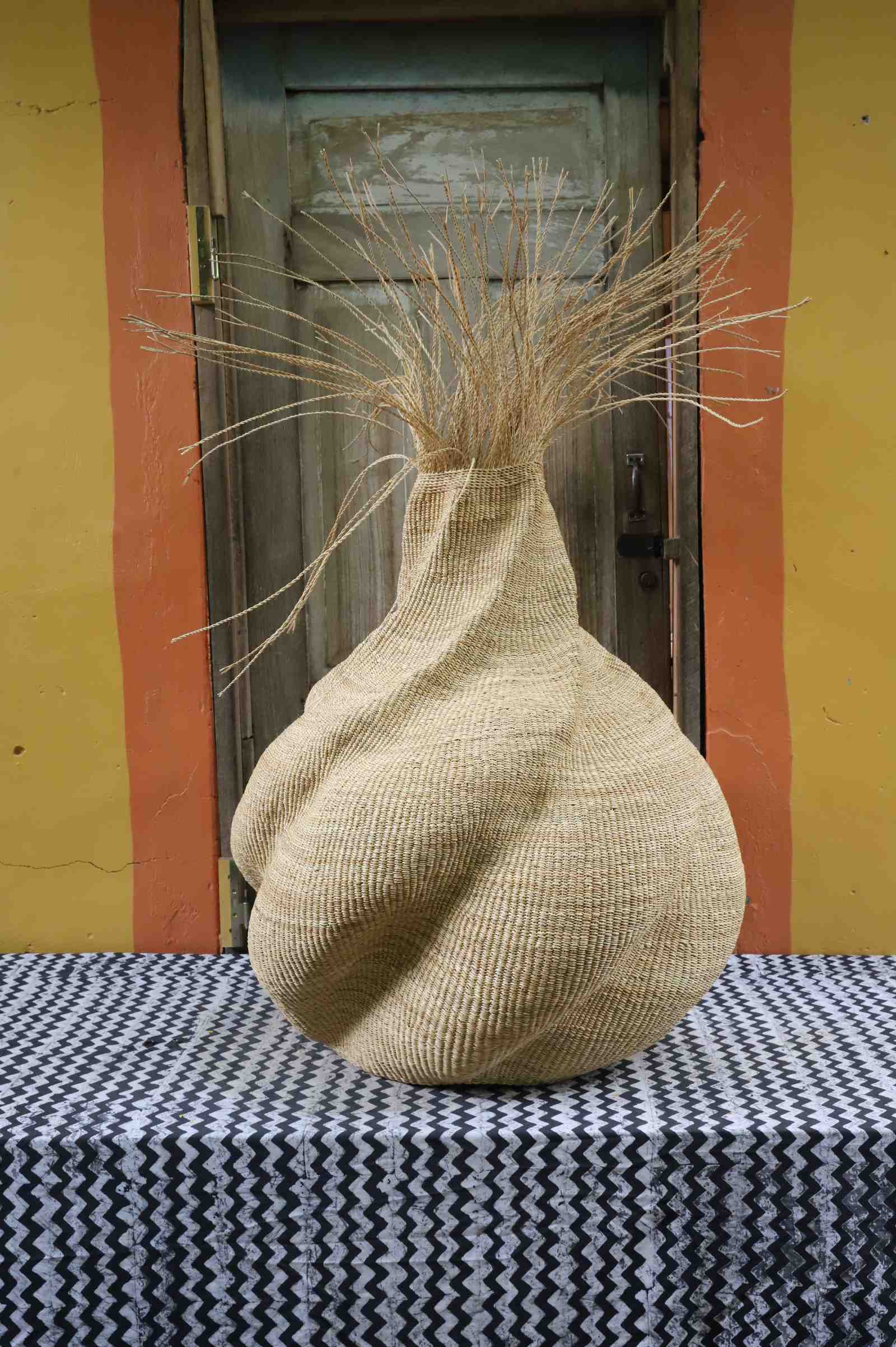 Handwoven art basket in natural straw with sculpted twisted form and dried grass at the top, on a chevron surface.