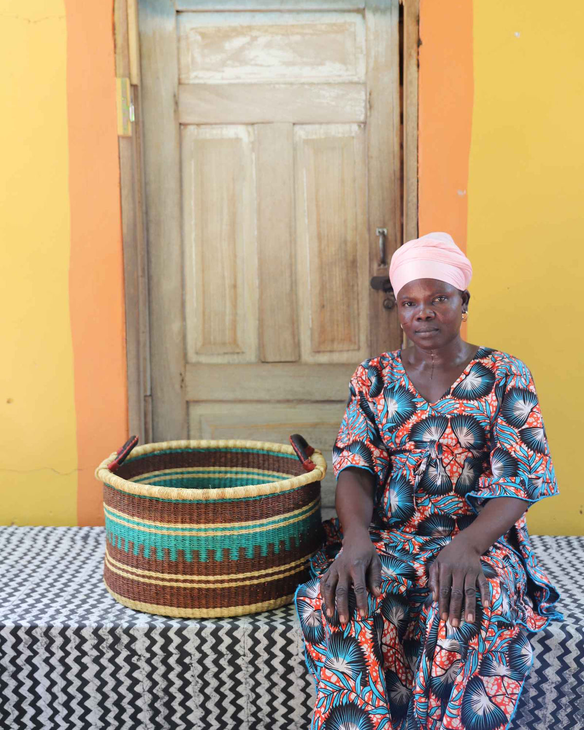 Medium handwoven Bolga storage tub in natural straw with dark banded stripes, styled beside a seated woman.