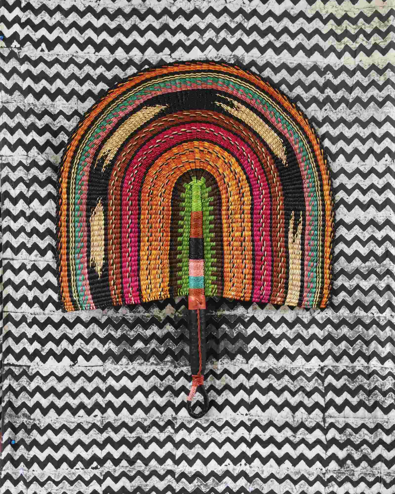 Naba Penga wall fan in natural straw with rust, green, pink and black rainbow arches, laid on a chevron cloth.