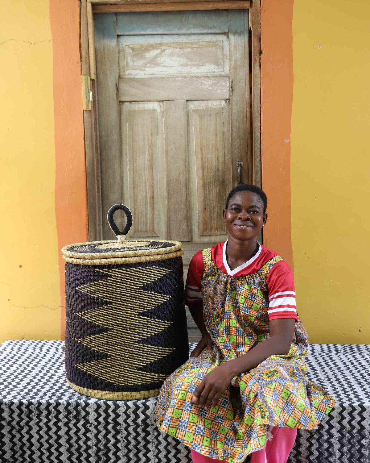 Handwoven Bolga laundry basket in natural with black zigzag band, beside a seated woman in a patterned dress.