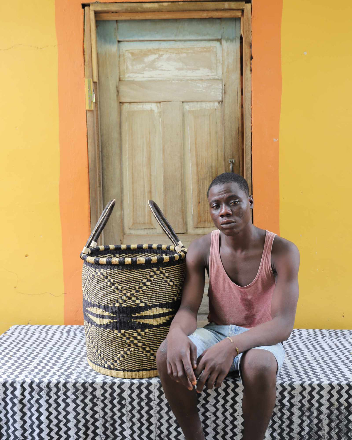 Medium handwoven Bolga laundry basket in natural with black wave motifs, beside a seated man in coral.