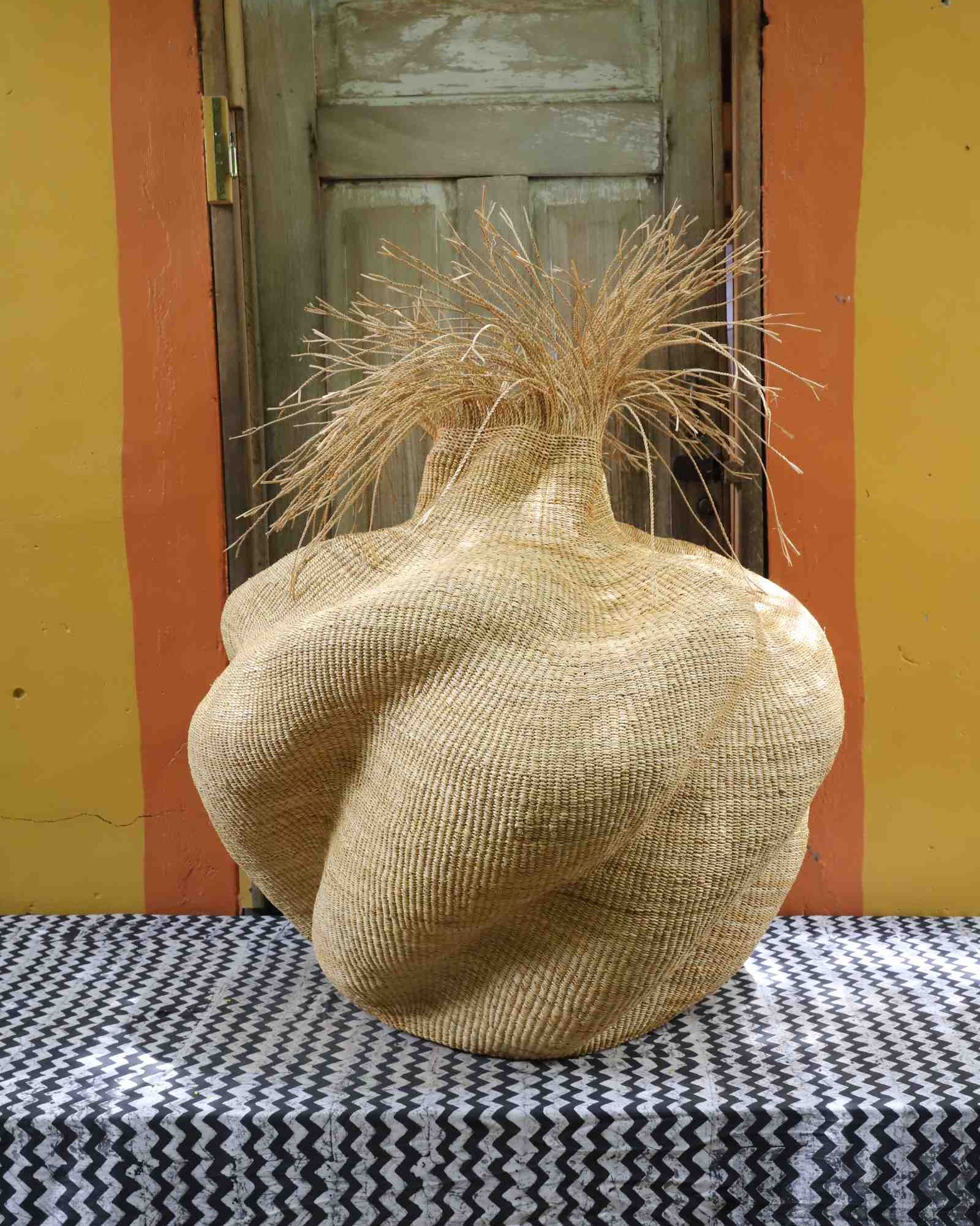 Side view of a handwoven natural straw art basket showing its organic lobed form and dried grass stems, on a chevron surface.