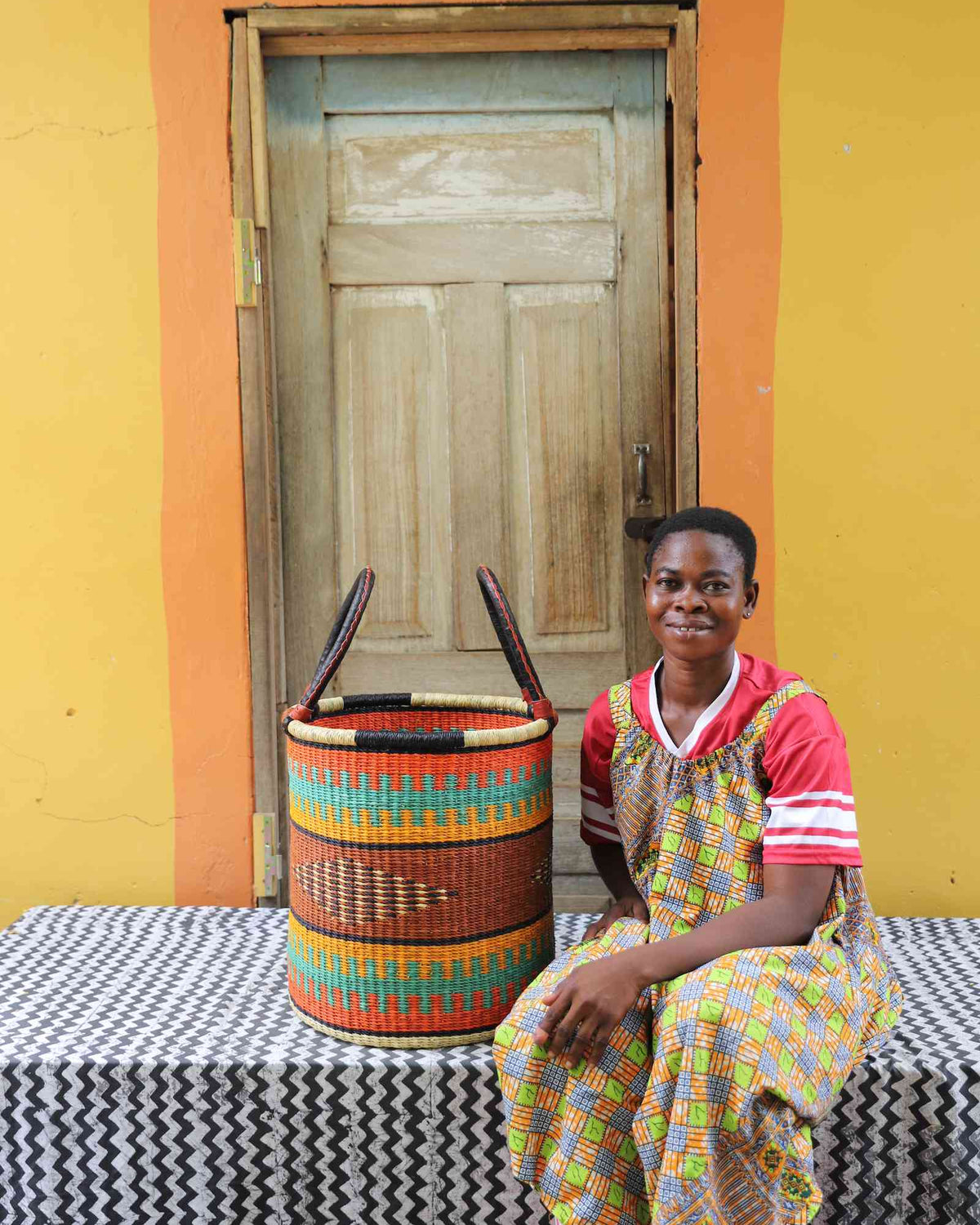 Small handwoven Bolga laundry basket in dyed orange with green and red bands, beside a seated woman.