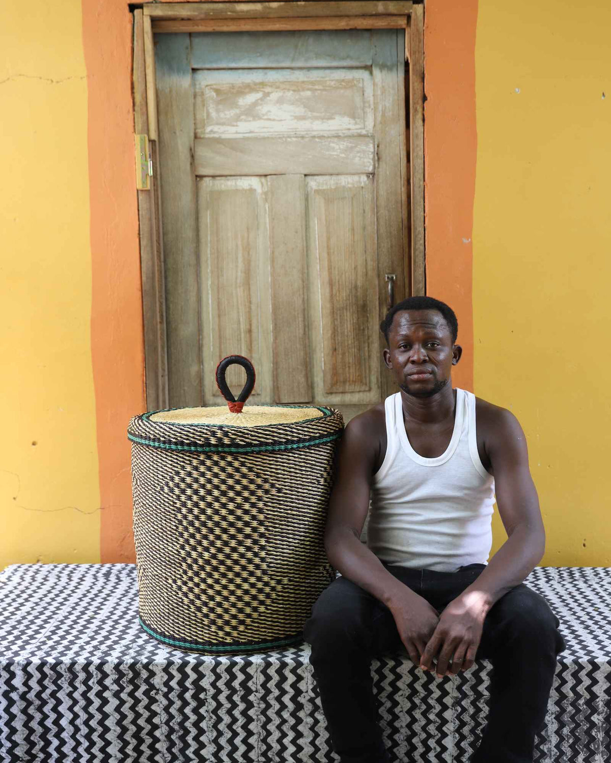 Handwoven Bolga laundry basket in natural straw with black checker pattern, beside a seated man in a white vest.