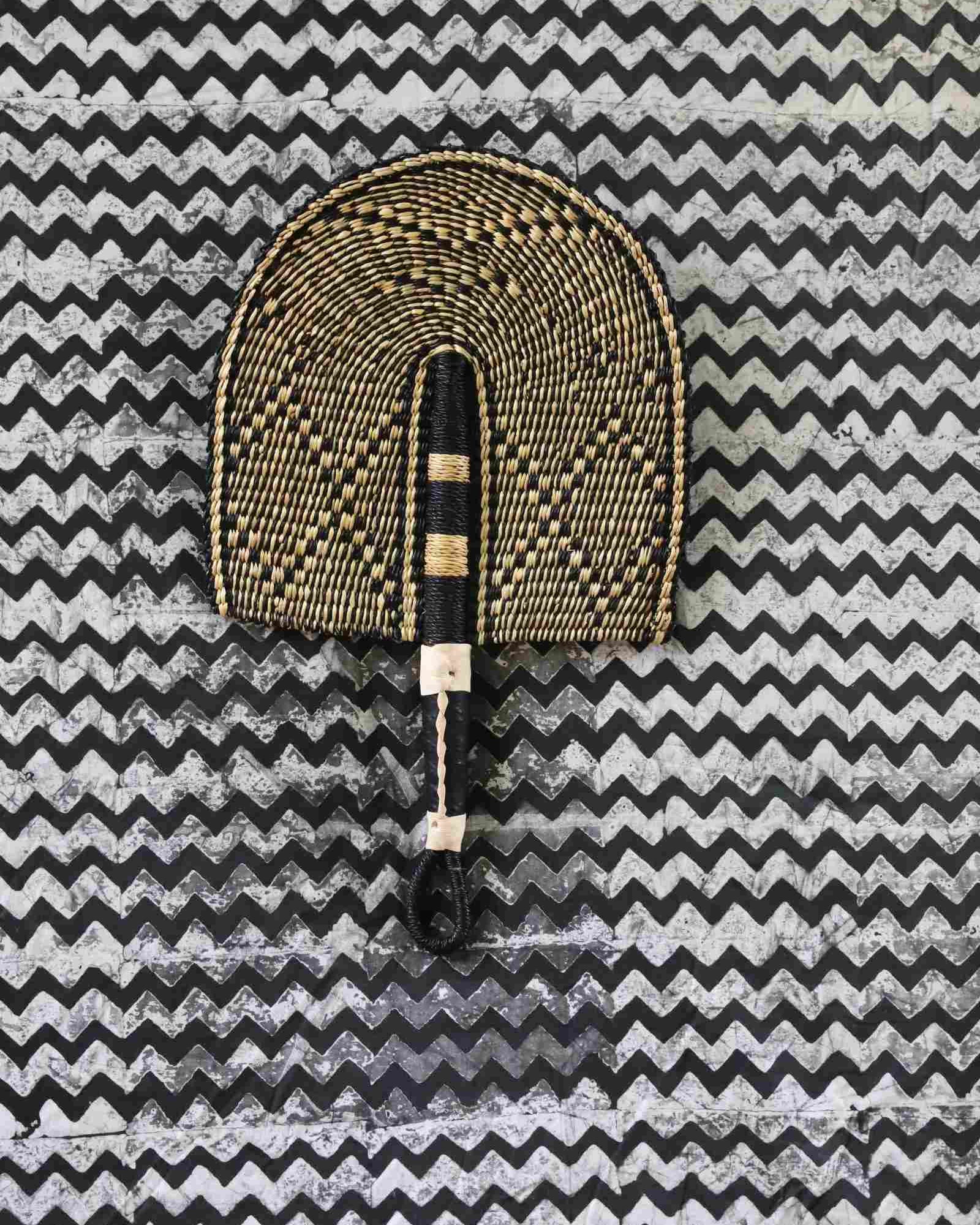 Bolga hand fan in natural straw with a dark chevron pattern and beaded handle, laid on a chevron cloth.
