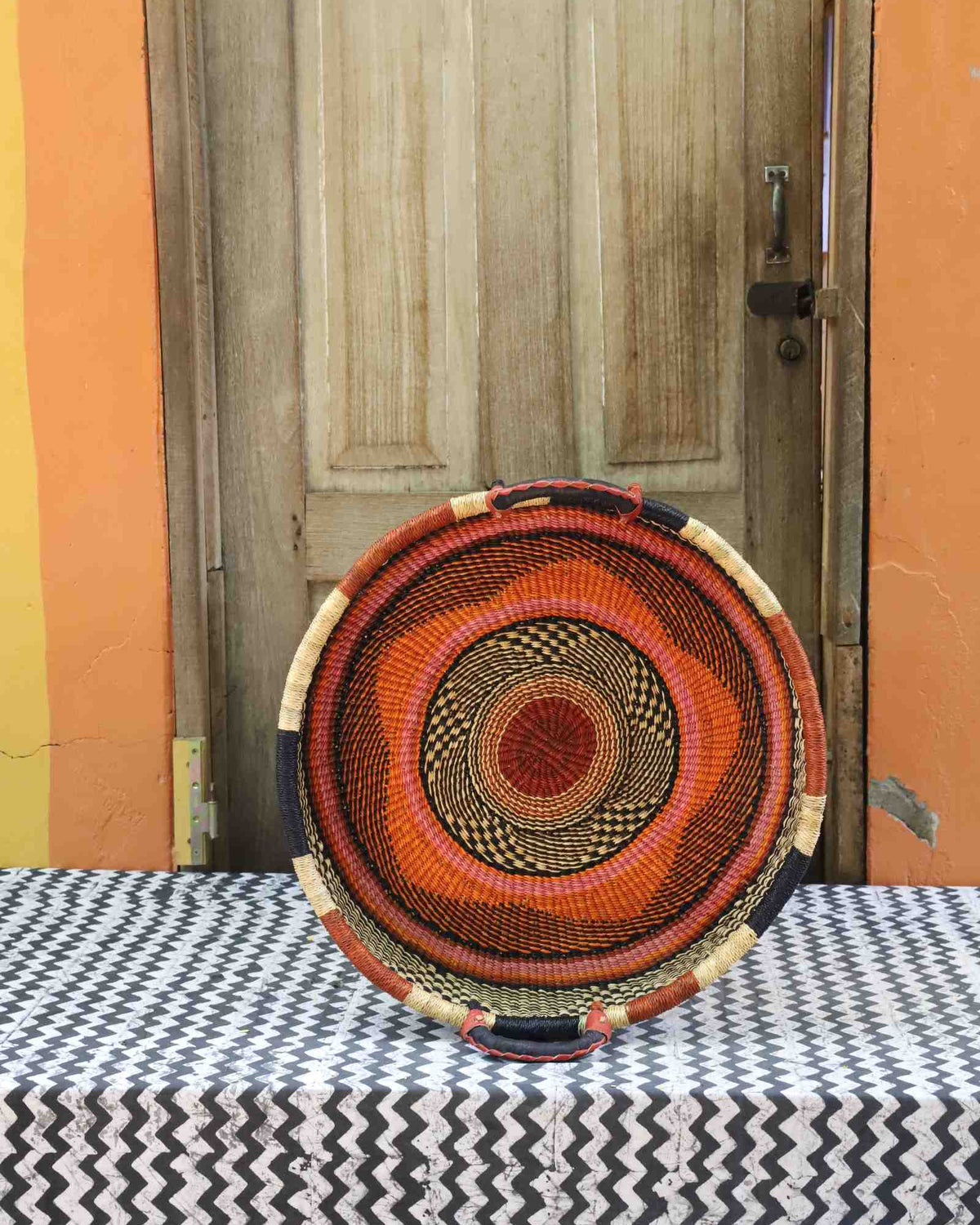 Handwoven Bolga wall tray disc in dyed rust-orange with concentric black ring pattern.