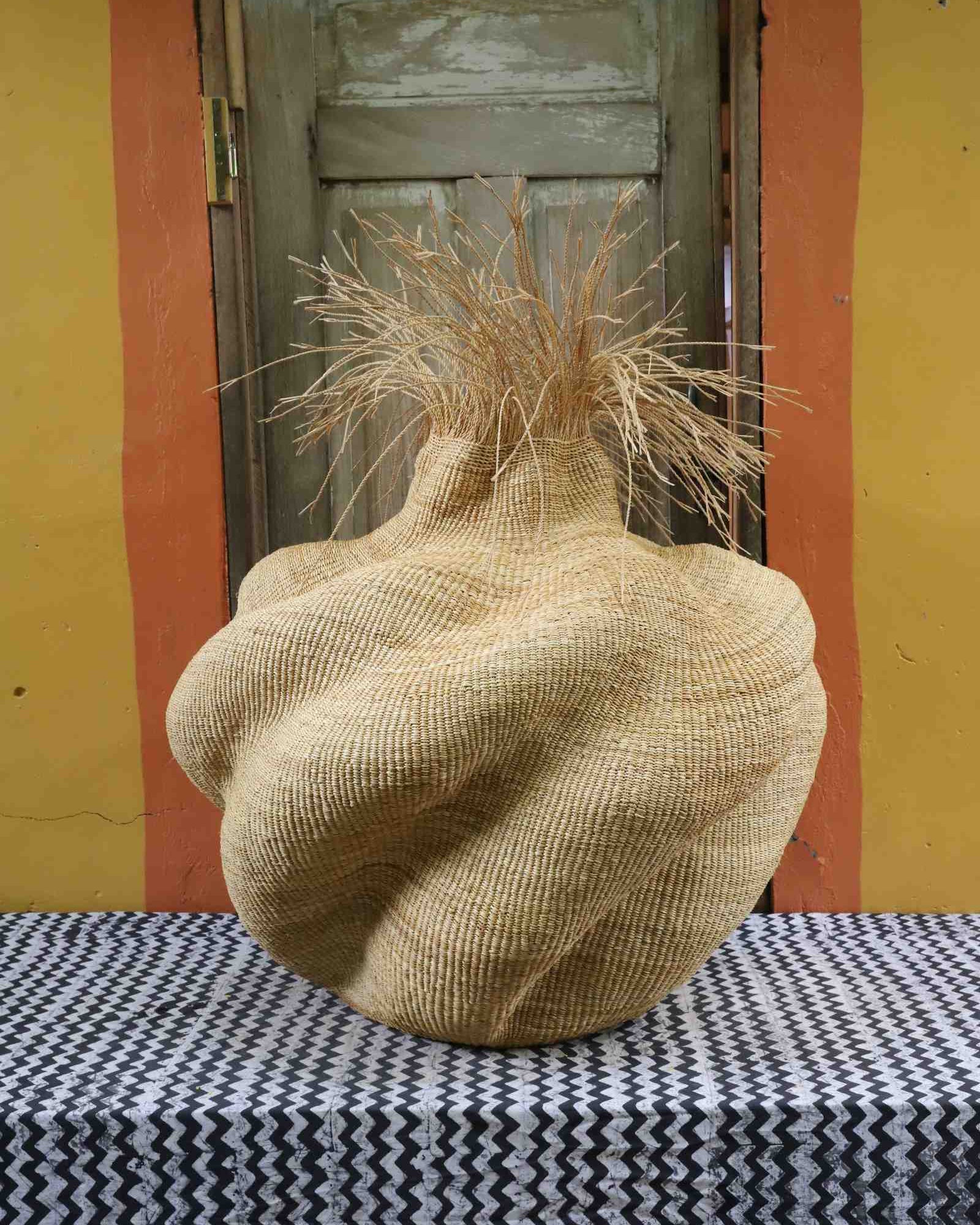 Handwoven art basket in natural straw with organic lobed body and dried grass stems at the top, on a chevron surface.