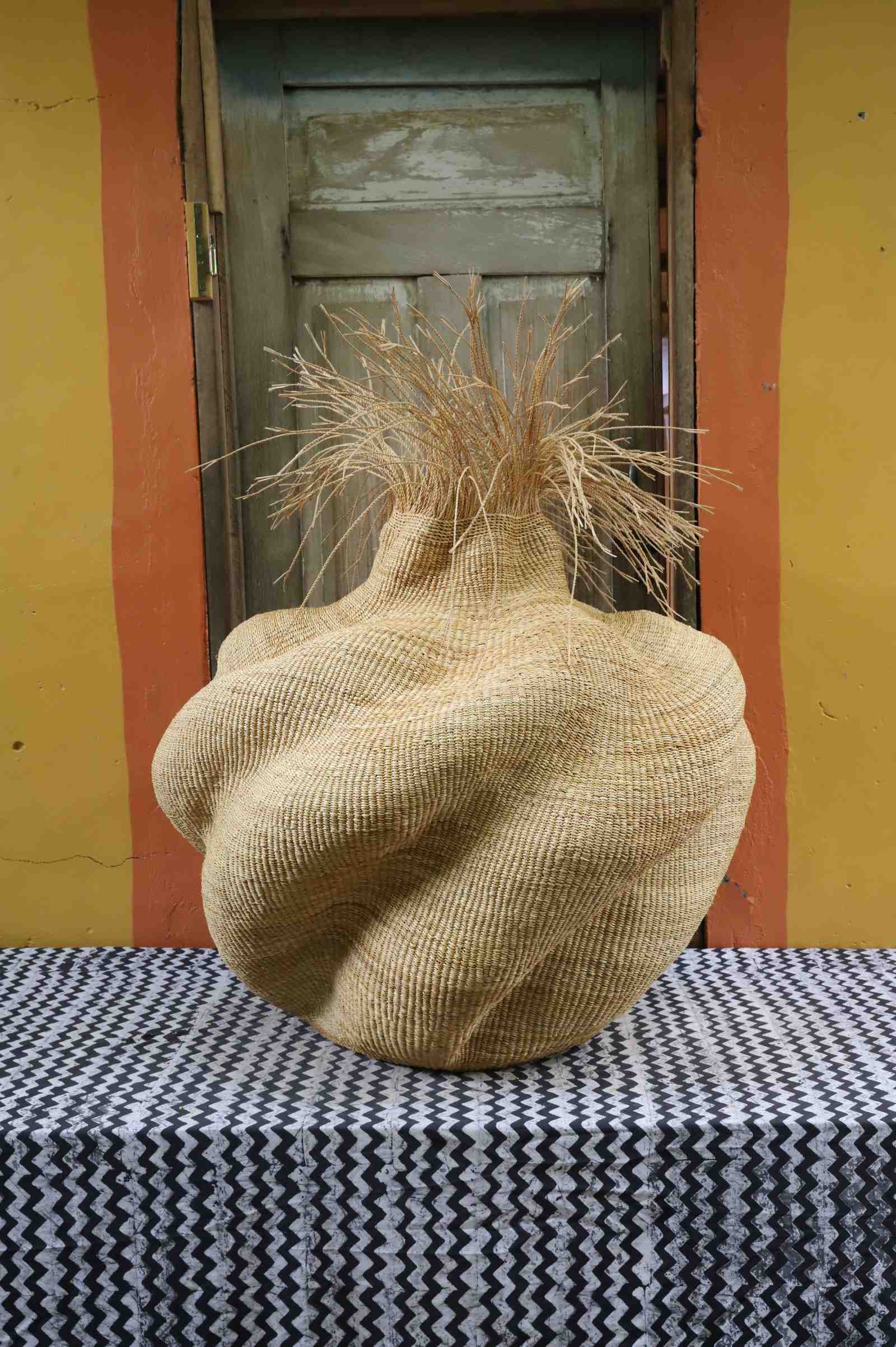 Handwoven art basket in natural straw with organic lobed body and dried grass stems at the top, on a chevron surface.
