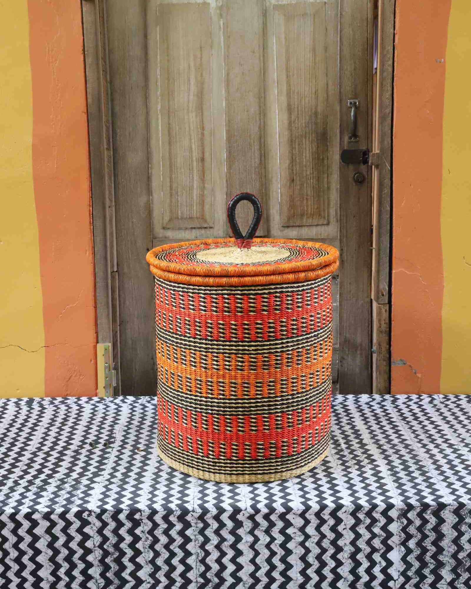 Coiled-rim lidded laundry basket in orange and black fine-striped weave, styled on a chevron cloth by a wooden door.