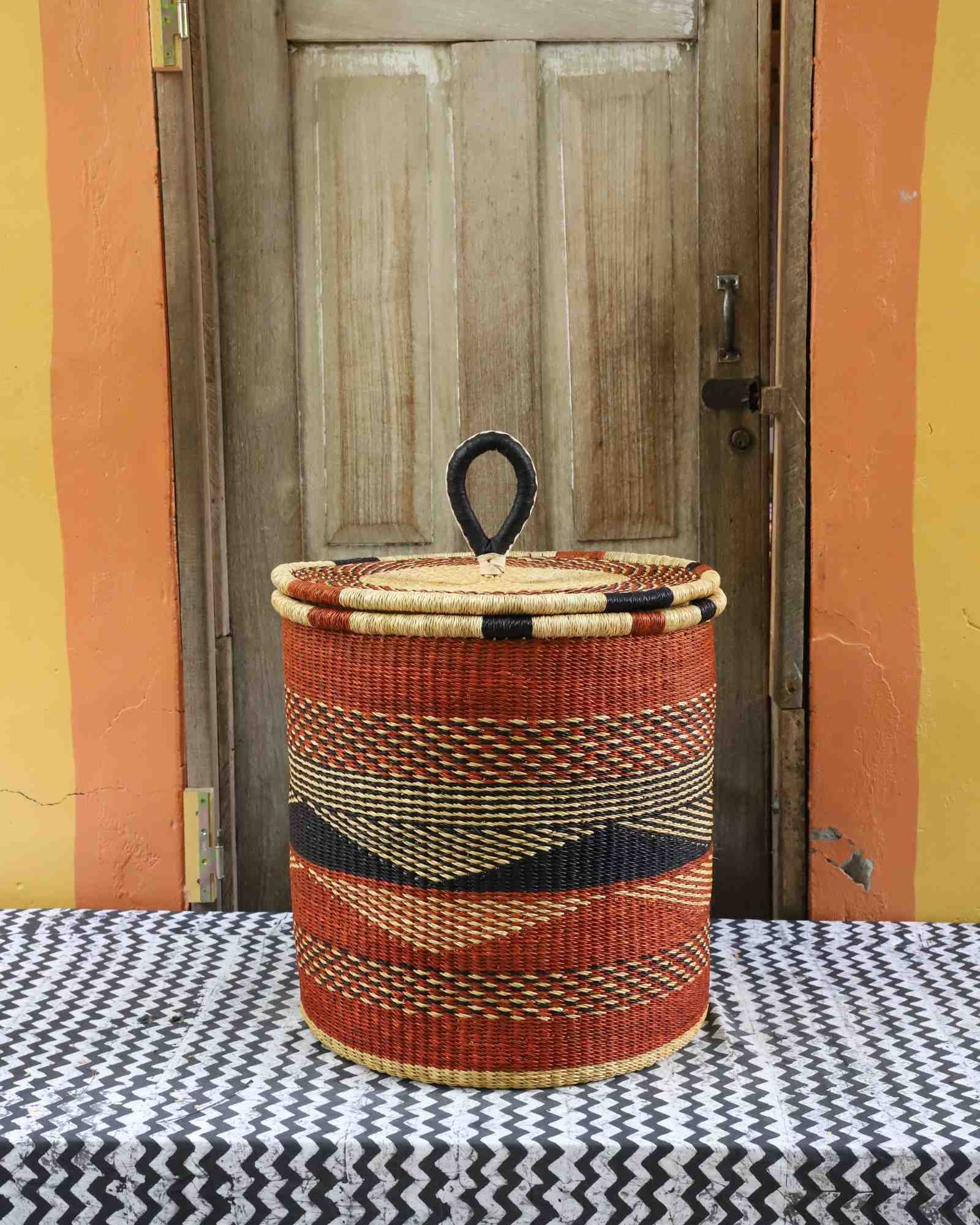 Handwoven Bolga laundry basket with coiled rim and lid in rust-orange with a dark wave motif and natural rim.