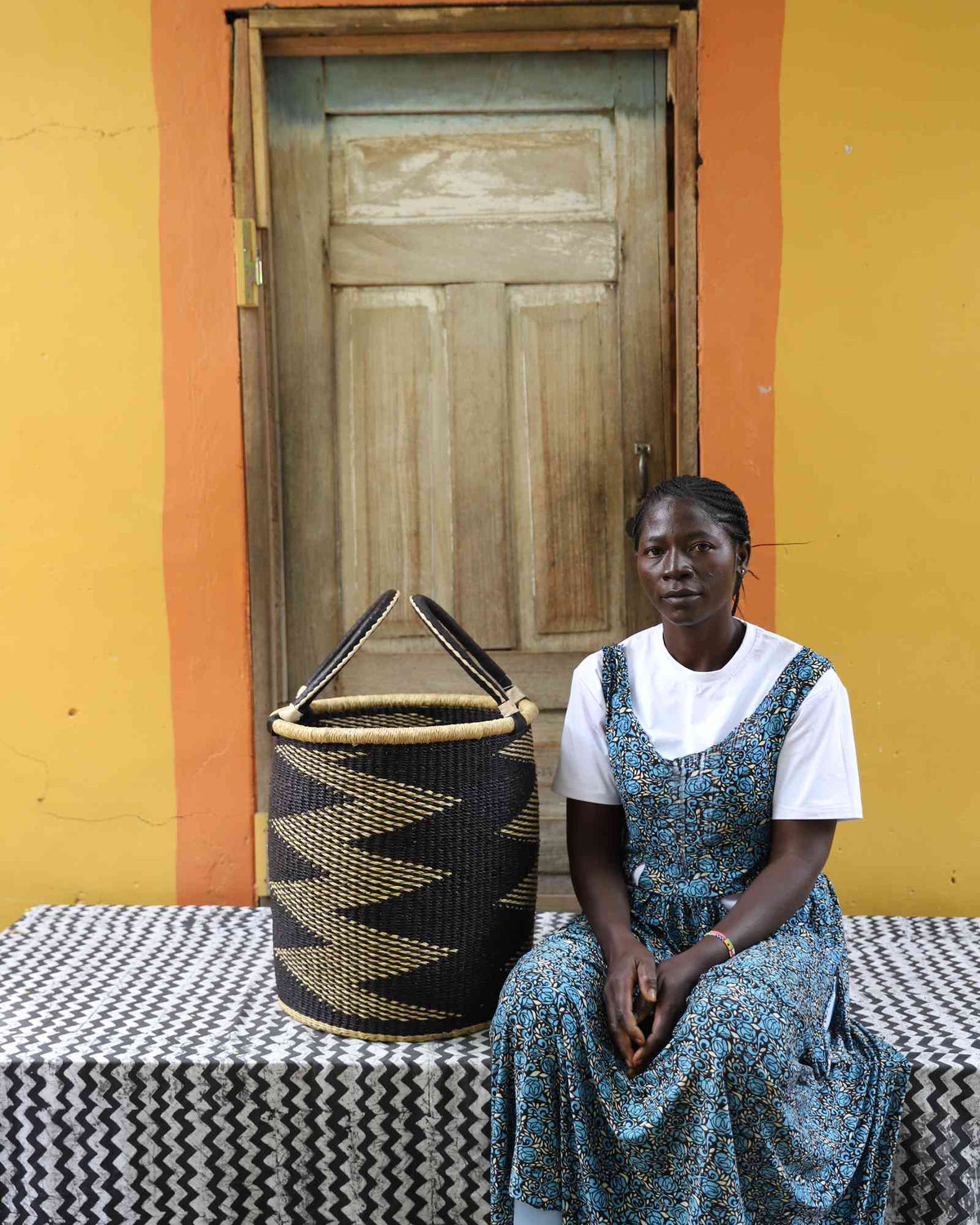 Small handwoven Bolga laundry basket in natural with bold black zigzag, beside a seated woman in green.