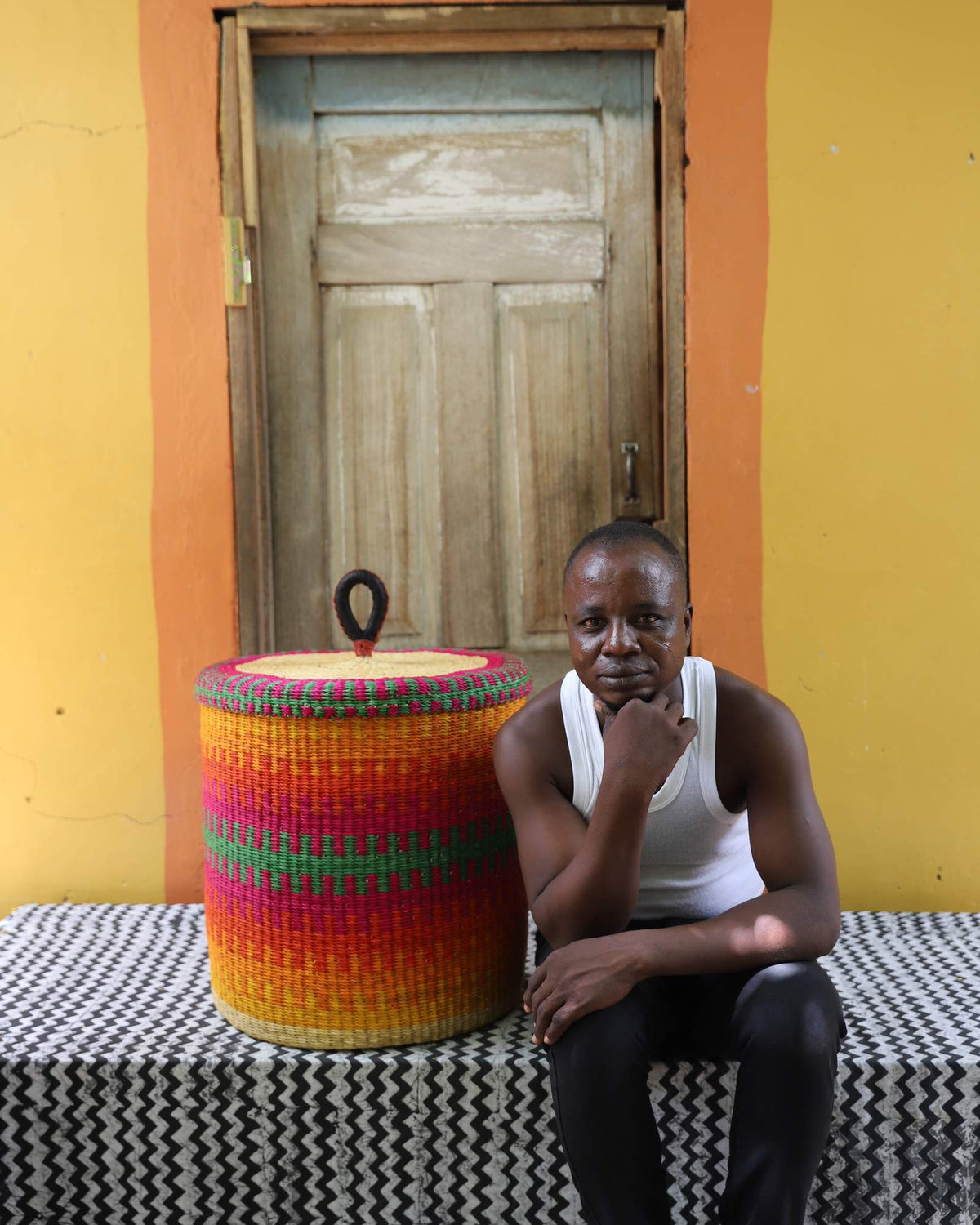 Handwoven Bolga laundry basket in fine multicolour stripes, beside a seated man in a white vest.