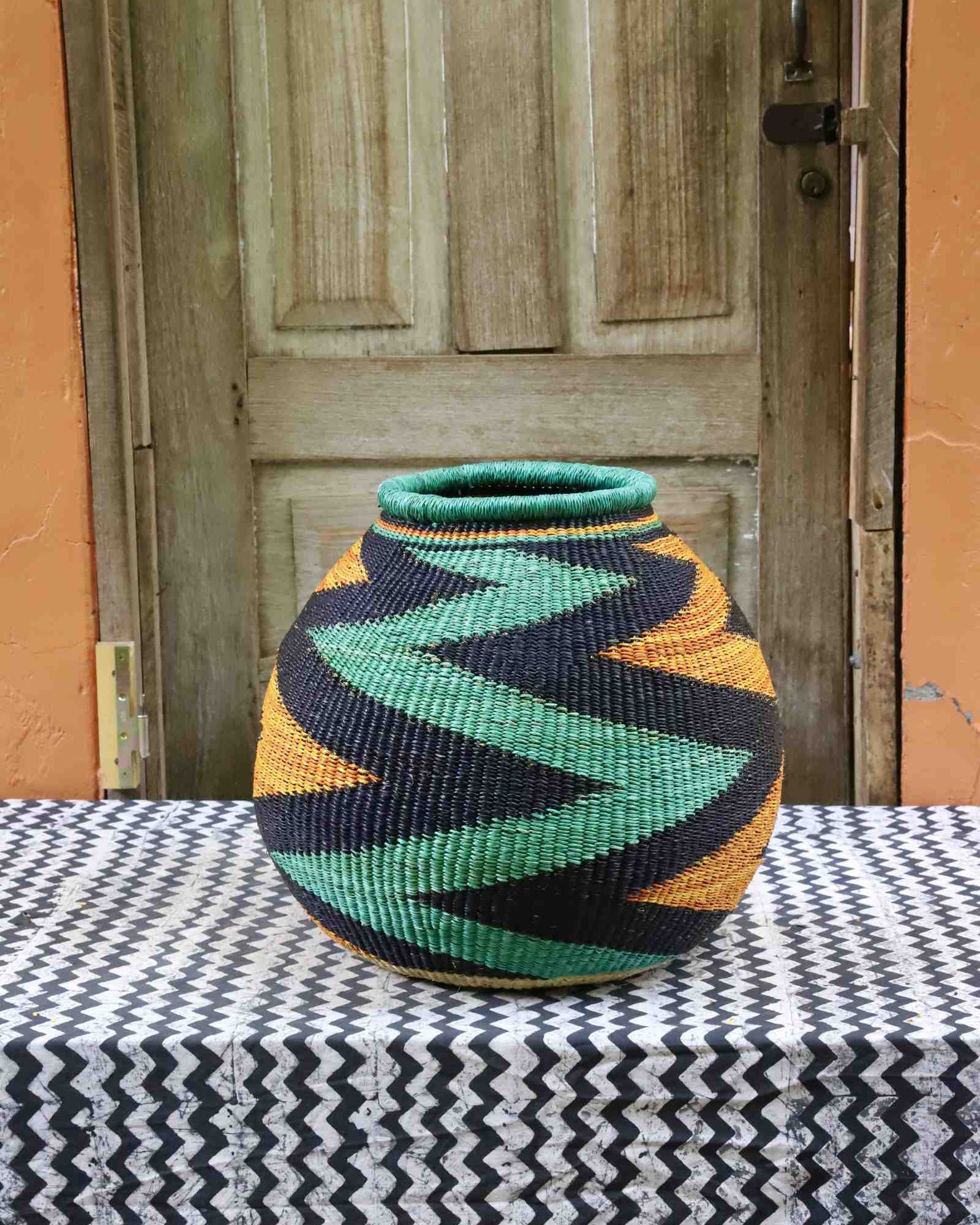 Handwoven African art basket with bold diagonal zigzag pattern in teal, black and amber, on a chevron surface.