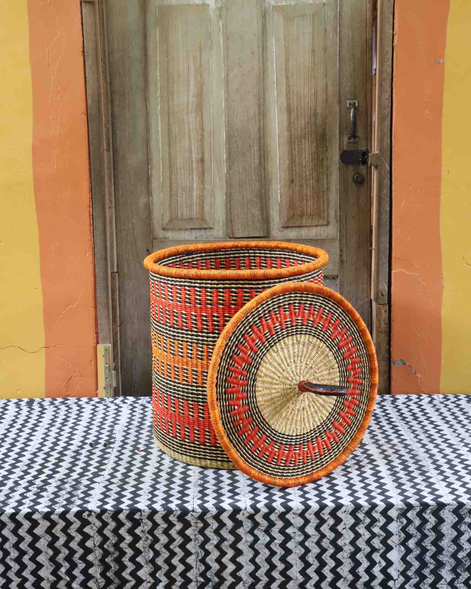Coiled-rim laundry basket in orange and black fine-striped weave, lid resting alongside on a chevron cloth.