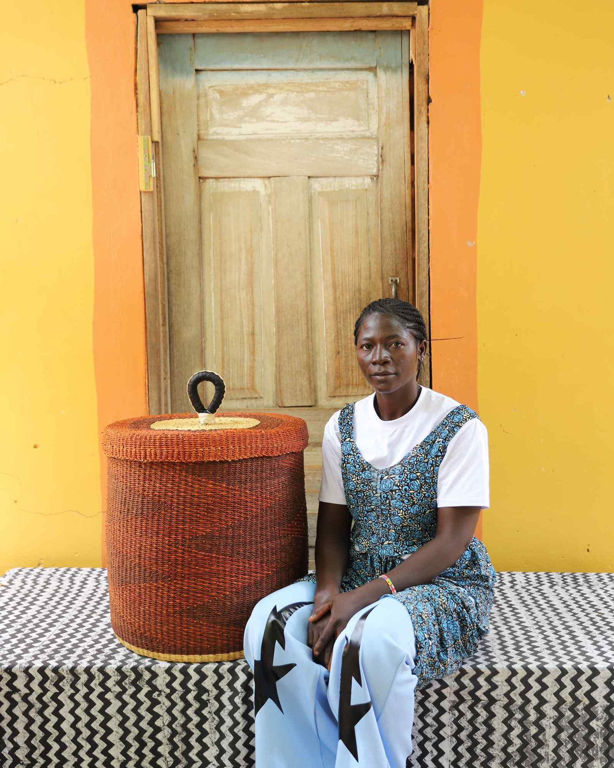 Handwoven Bolga laundry basket in dyed rust-red, beside a seated woman in blue and white.